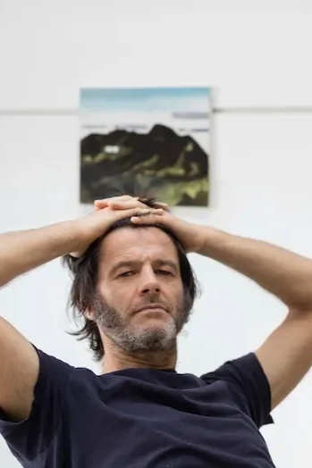 Man with dark hair and beard, wearing a black shirt, holding his head with both hands and looking at the camera, with blurred landscape picture hanging on the wall behind him.