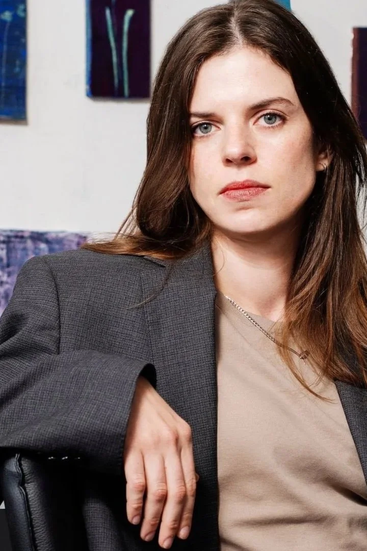 A woman with long brown hair and blue eyes wearing a gray blazer and beige top, sitting with her arm resting on the back of a chair, looking serious, with abstract paintings in the background.