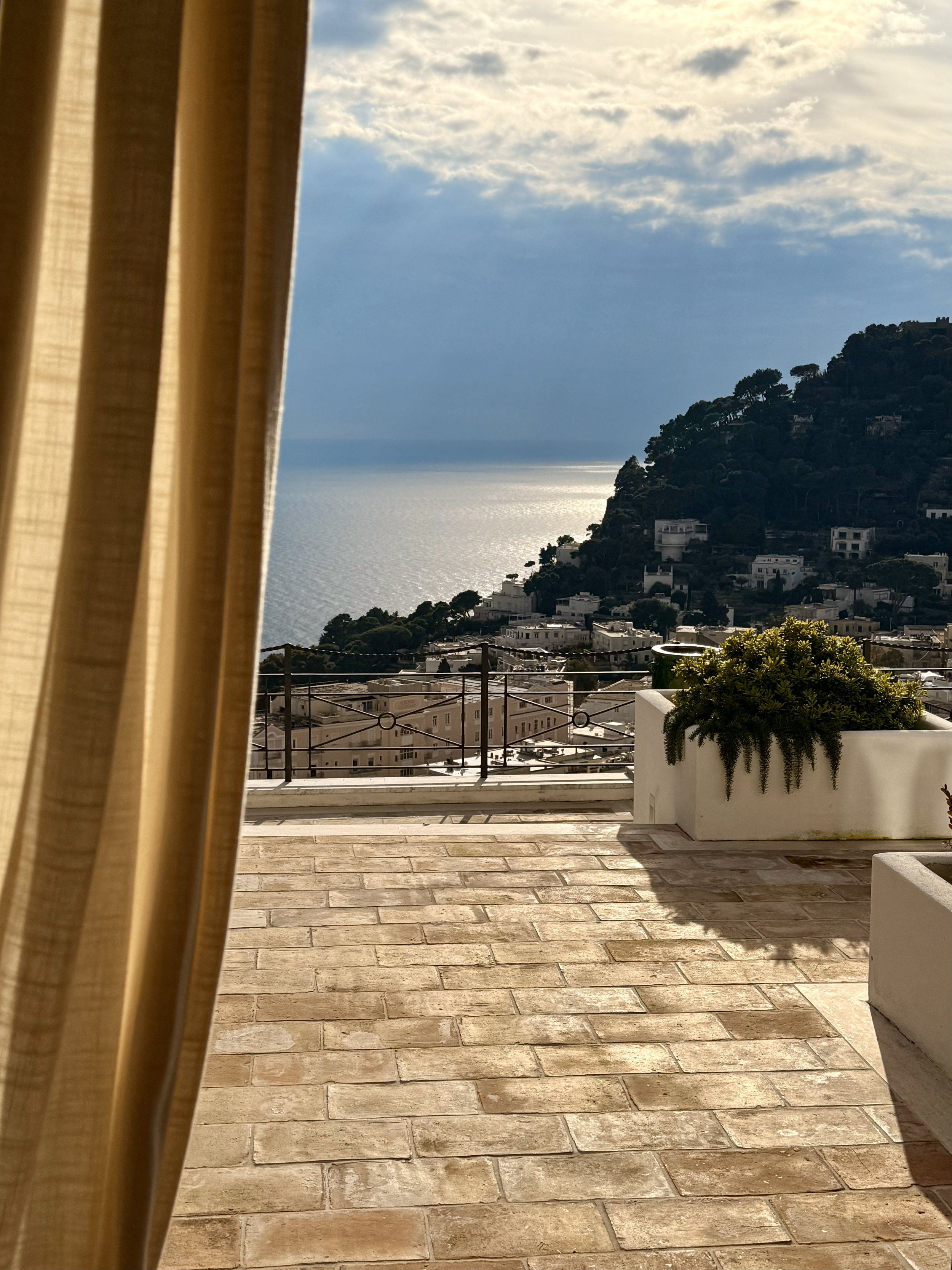 A balcony with a tiled floor and white planters with greenery, overlooking a hilly coastline with white buildings, a body of water, and a partly cloudy sky.