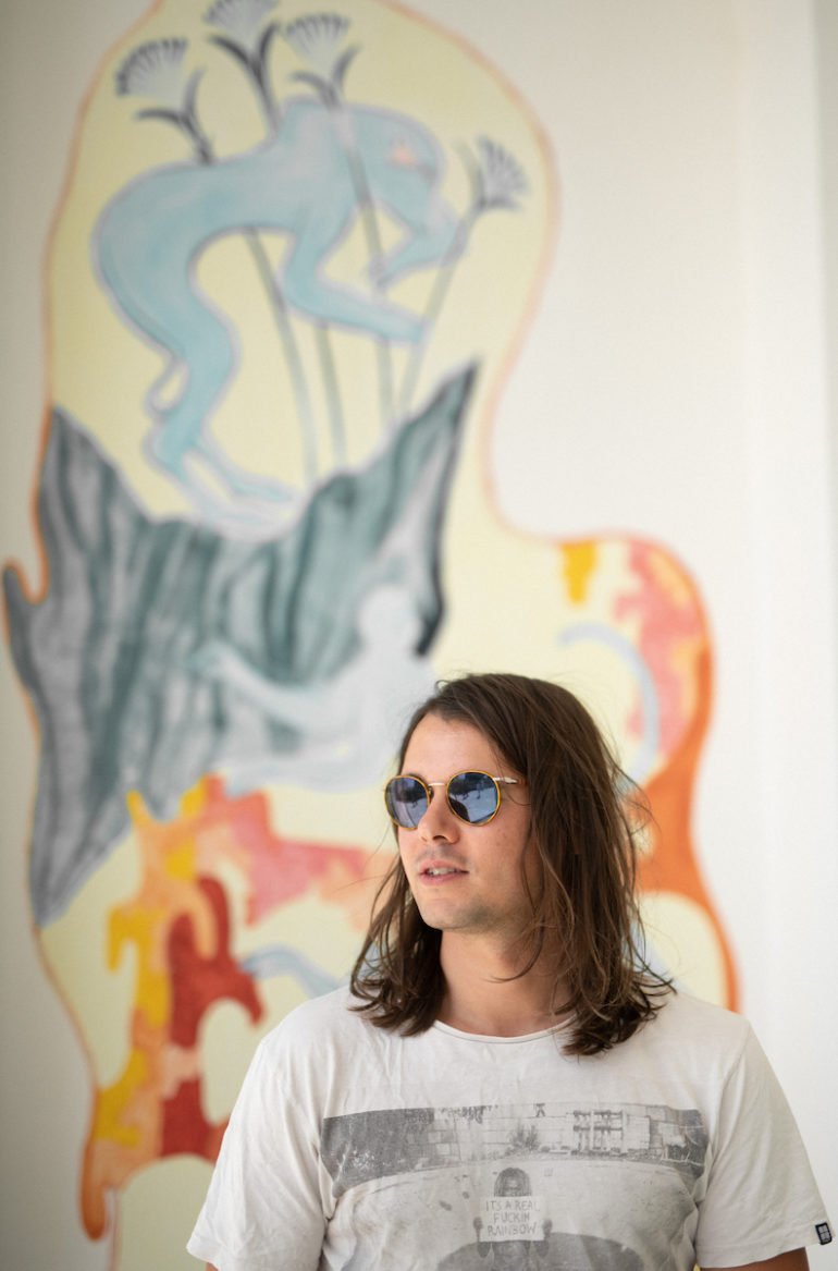 A young man with long brown hair wearing sunglasses and a white t-shirt, standing in front of a colorful graffiti art mural on a wall.