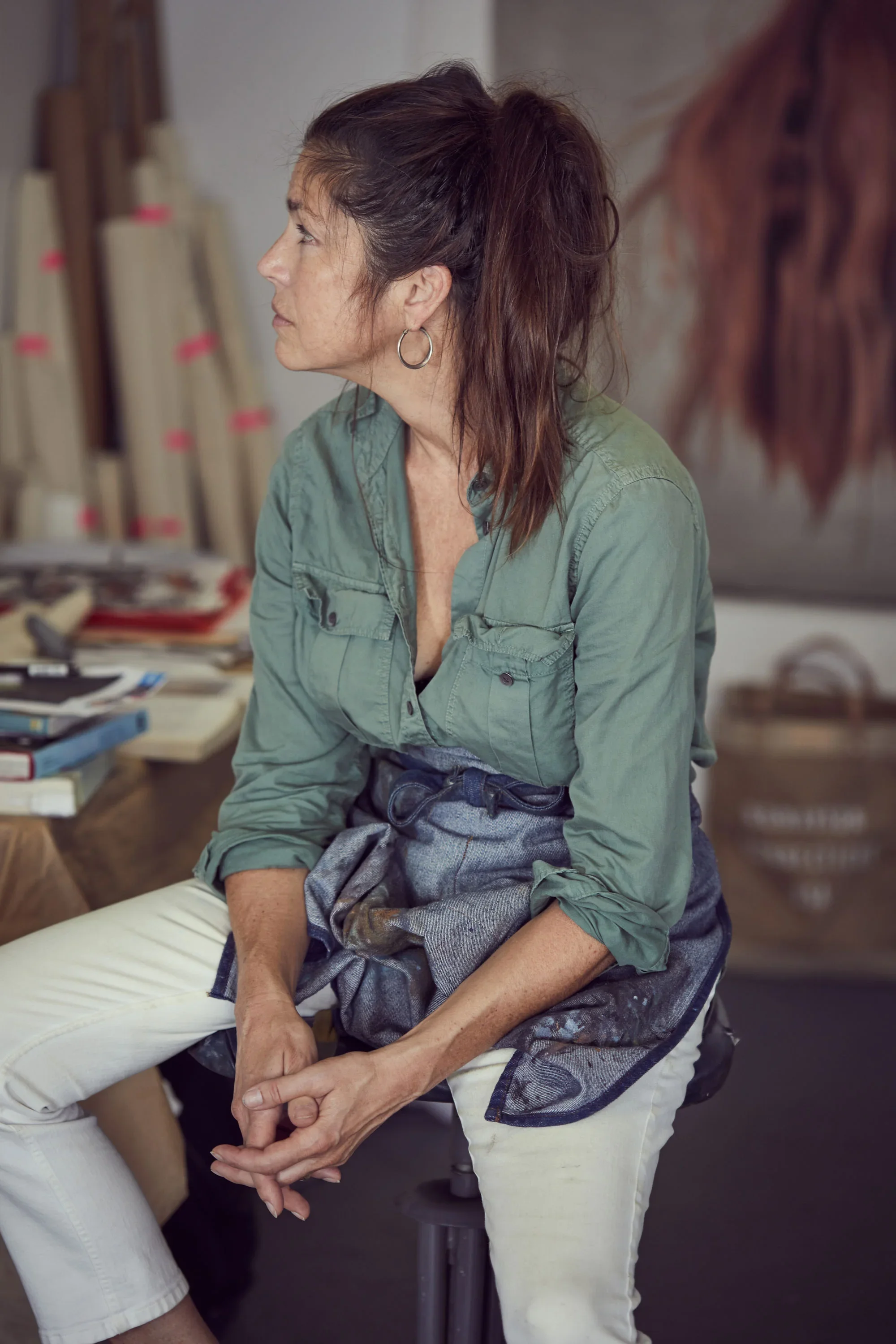 A woman with dark brown hair tied back, wearing hoop earrings and a green shirt, sitting in a studio or workshop with art supplies and paintings in the background.