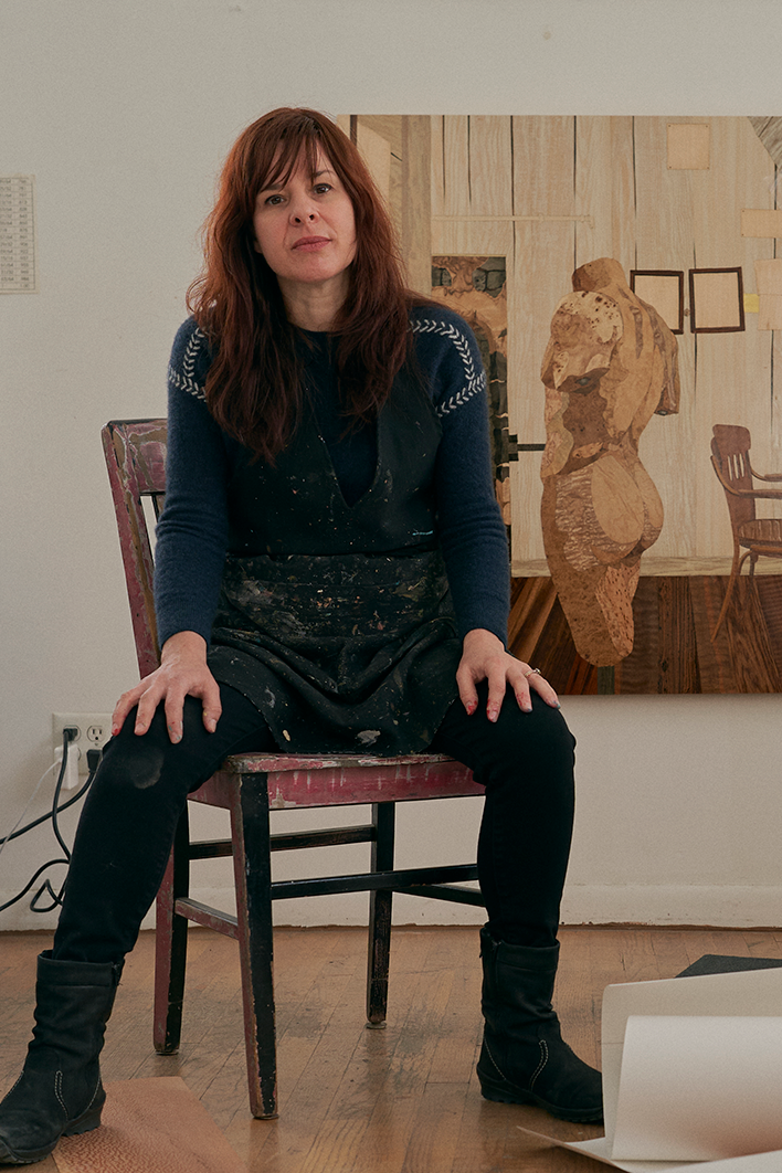 Woman sitting on a wooden chair in an art studio, with a large wooden abstract sculpture and art pieces in the background.