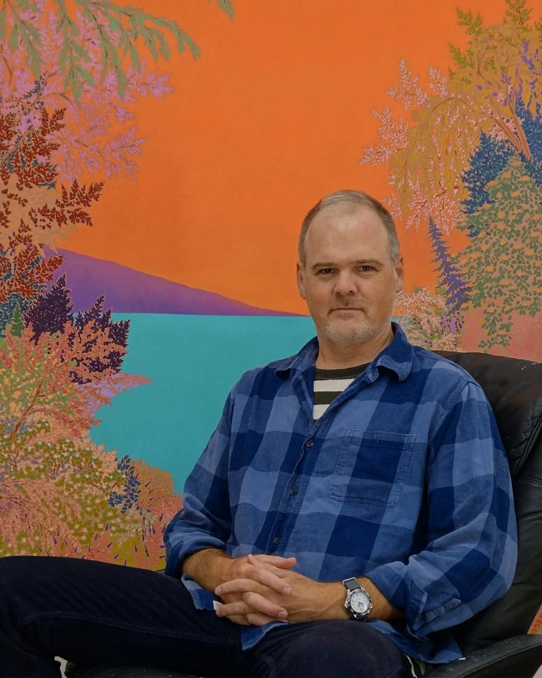 A man with short hair, wearing a blue plaid shirt, sitting on a black chair with his hands clasped, in front of a colorful abstract landscape mural with trees and a body of water in vibrant colors.