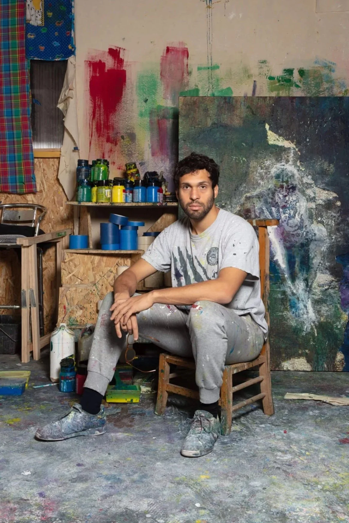 A man with dark hair and beard sitting on a small wooden chair in an artist's studio, surrounded by colorful paint jars and abstract paintings on the walls.