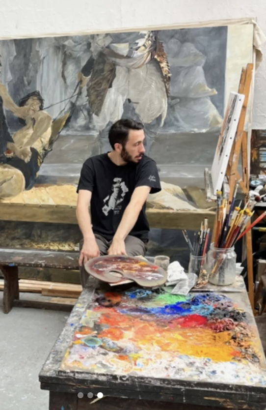 An artist sitting at a cluttered table with colorful paint splatters, holding a paint palette, in front of a large, partially finished mural with figures in a landscape.