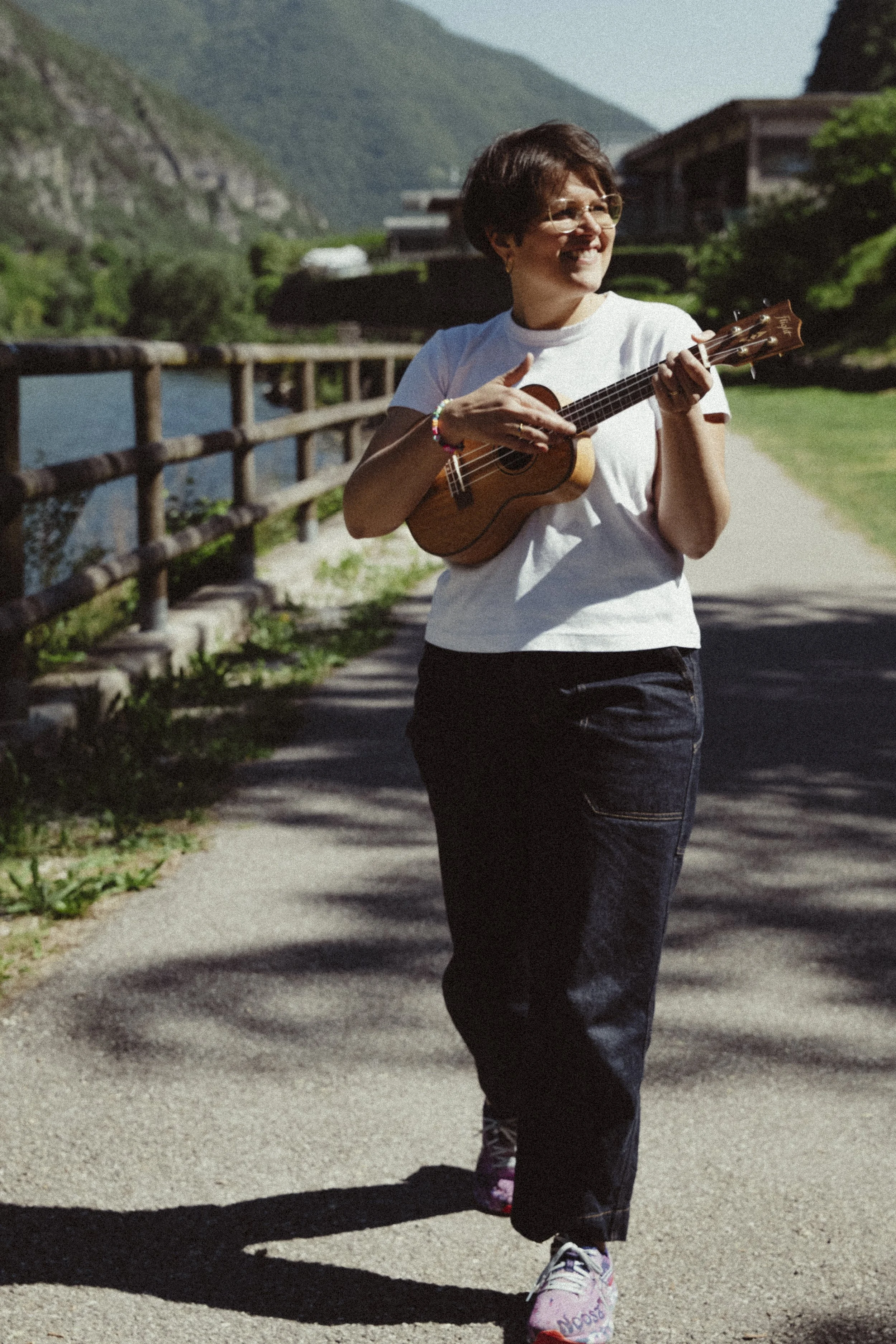 Una donna che cammina lungo un sentiero con un clima soleggiato, su un ponte di legno, che suona una chitarra e indossa occhiali, pantaloni neri e scarpe da ginnastica.