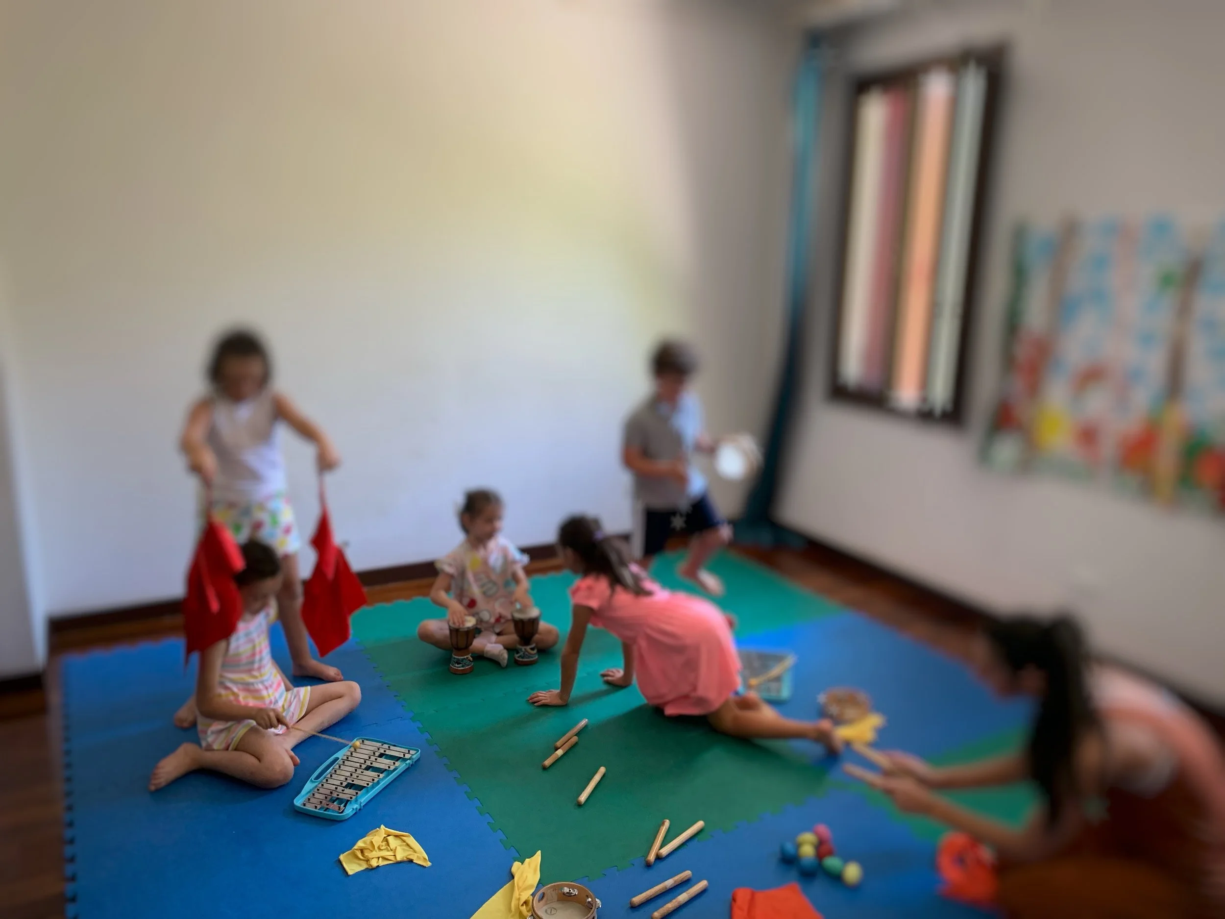 Bambini che partecipano a una lezione di musica in una stanza con pareti bianche, seduti su tappetini colorati, con strumenti musicali come xilofoni e tamburi. Insegnante che guida l'attività.