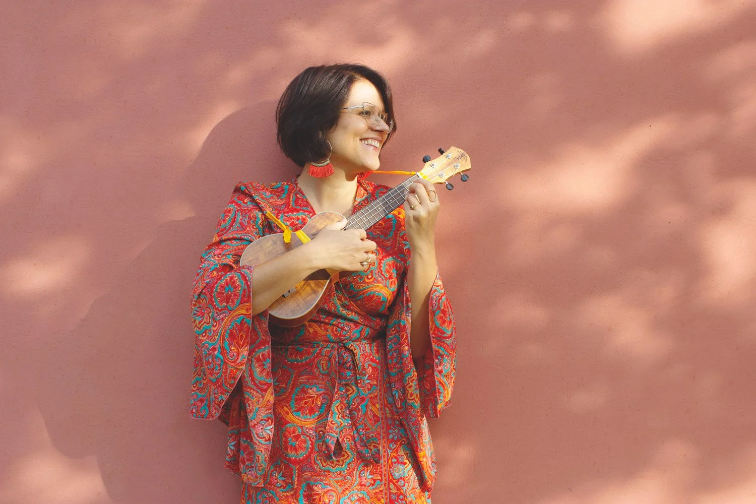 Una donna sorridente che suona un ukulele, indossa un abito colorato con motivi floreali, si trova davanti a una parete rosa.