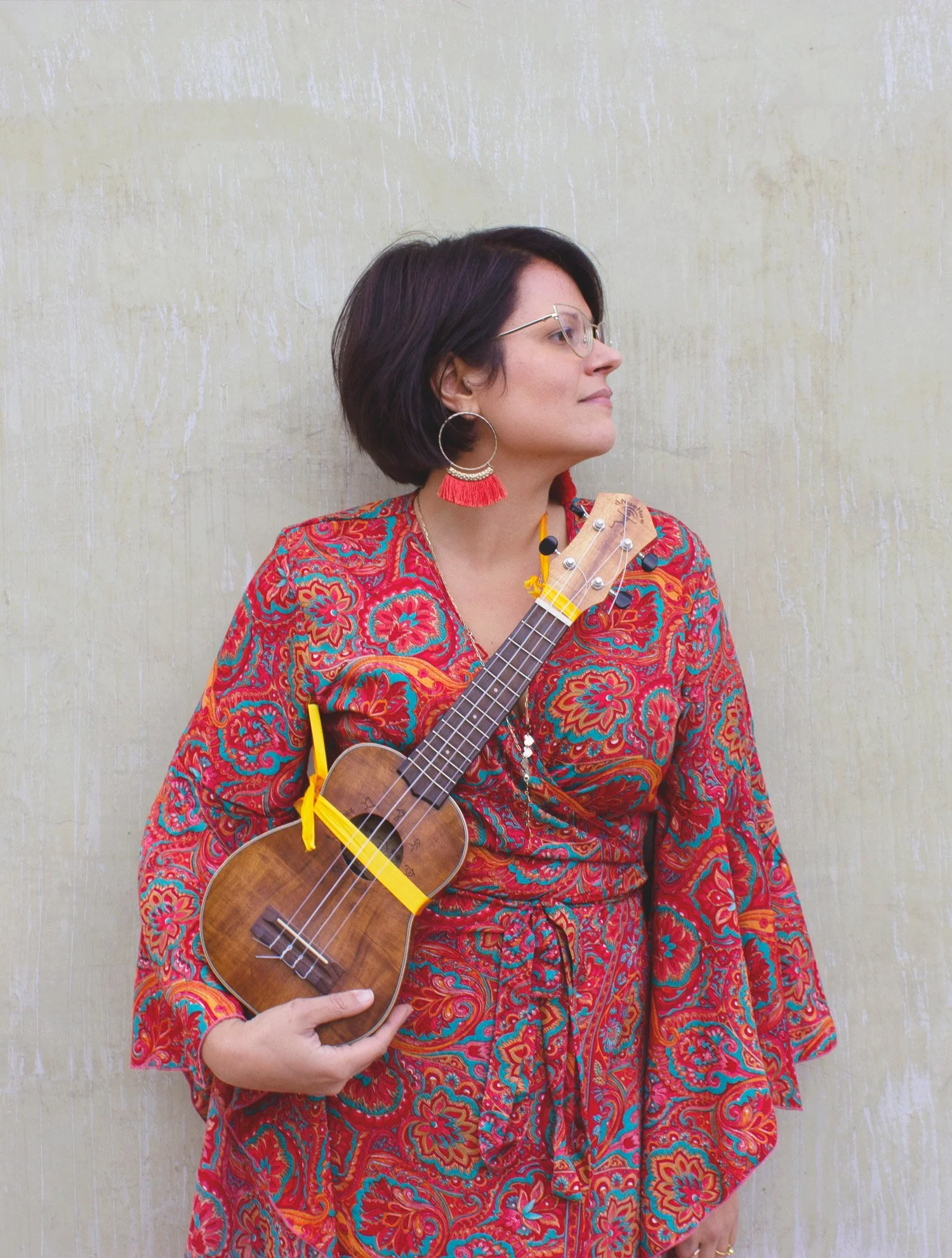 Donna con capelli corti e occhiali, indossa orecchini grandi con nappe rosse e abito con motivo floreale rosso e blu, tiene un ukulele e sta contro una parete beige.