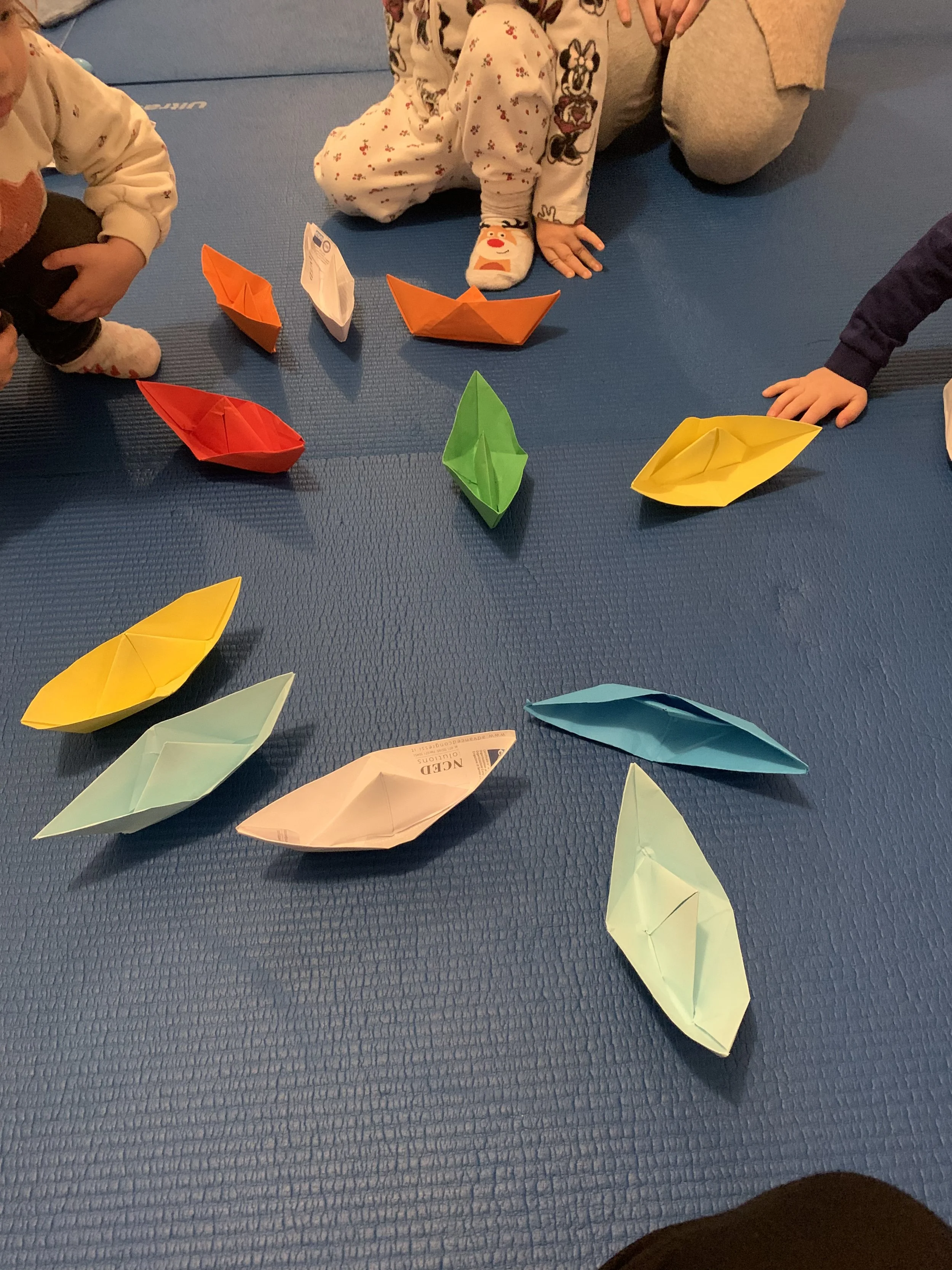 Bambini in piedi e in ginocchio intorno a barche di carta colorate sul pavimento, in un'ambiente interno.