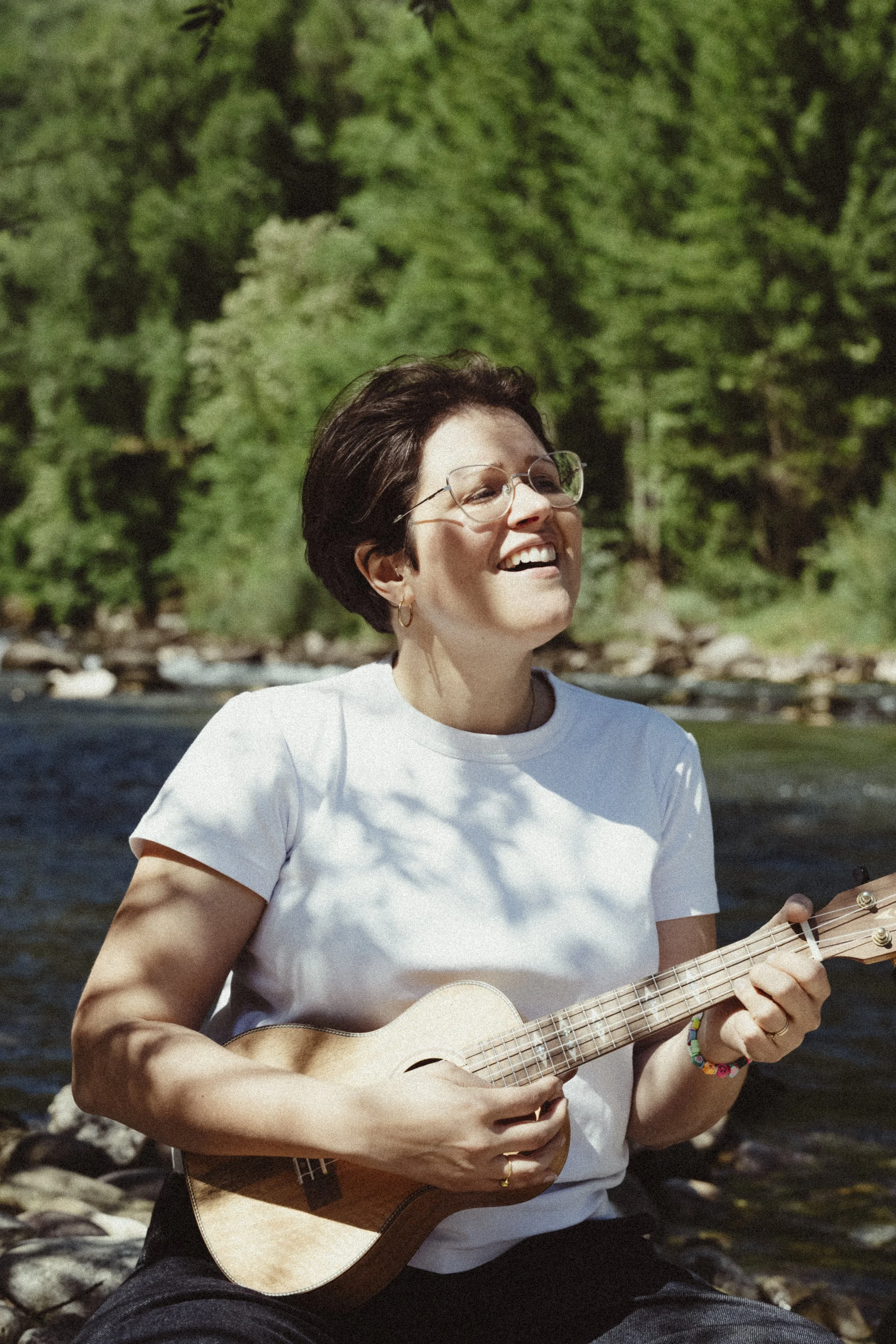 Una donna con capelli corti, occhiali, che sorride mentre suona una chitarra acustica all'aperto vicino a un fiume e alla natura
