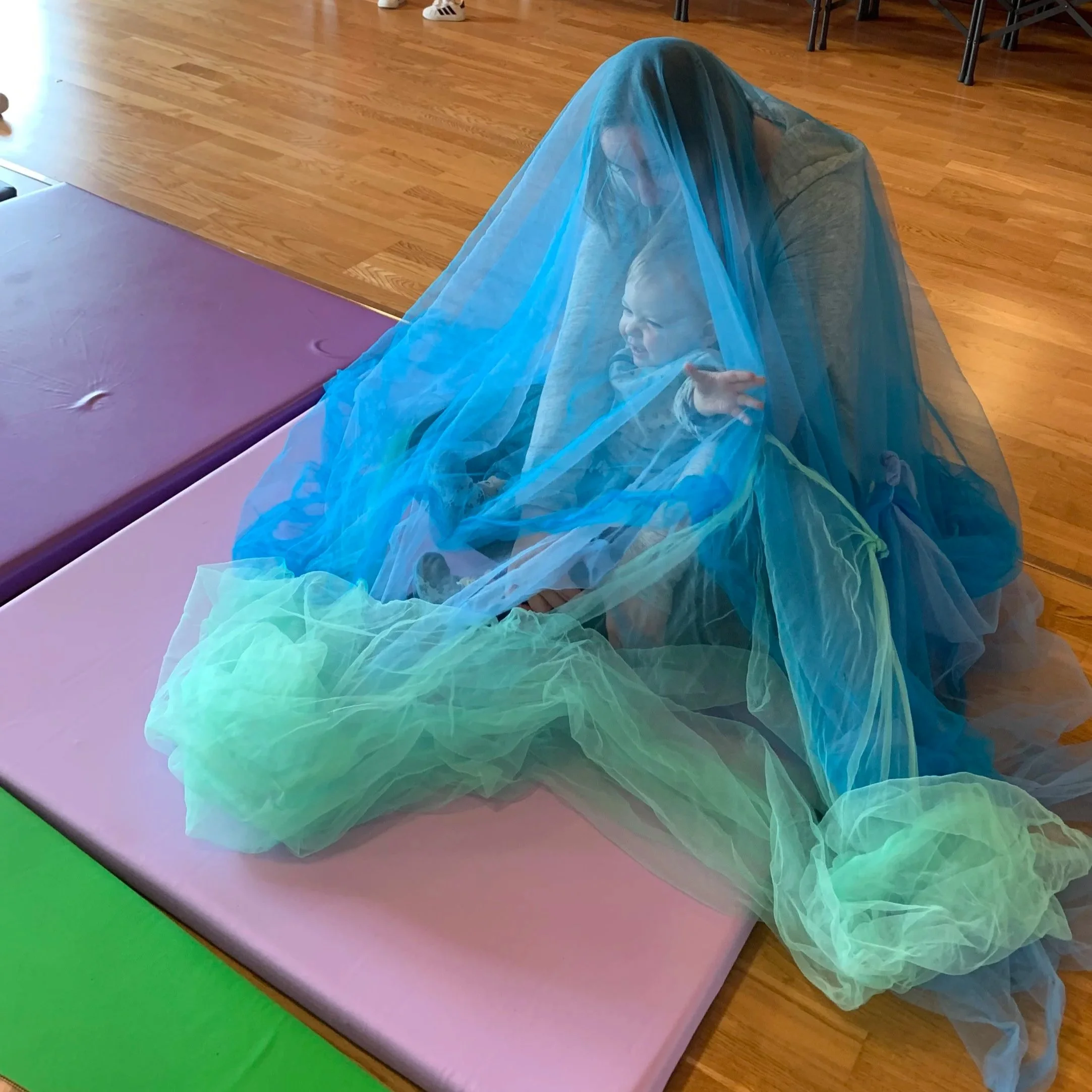 Bambino felice coperto da una tenda di tessuto trasparente azzurro, seduto su una panca in un ambiente interno con pavimento in legno e pareti colorate.