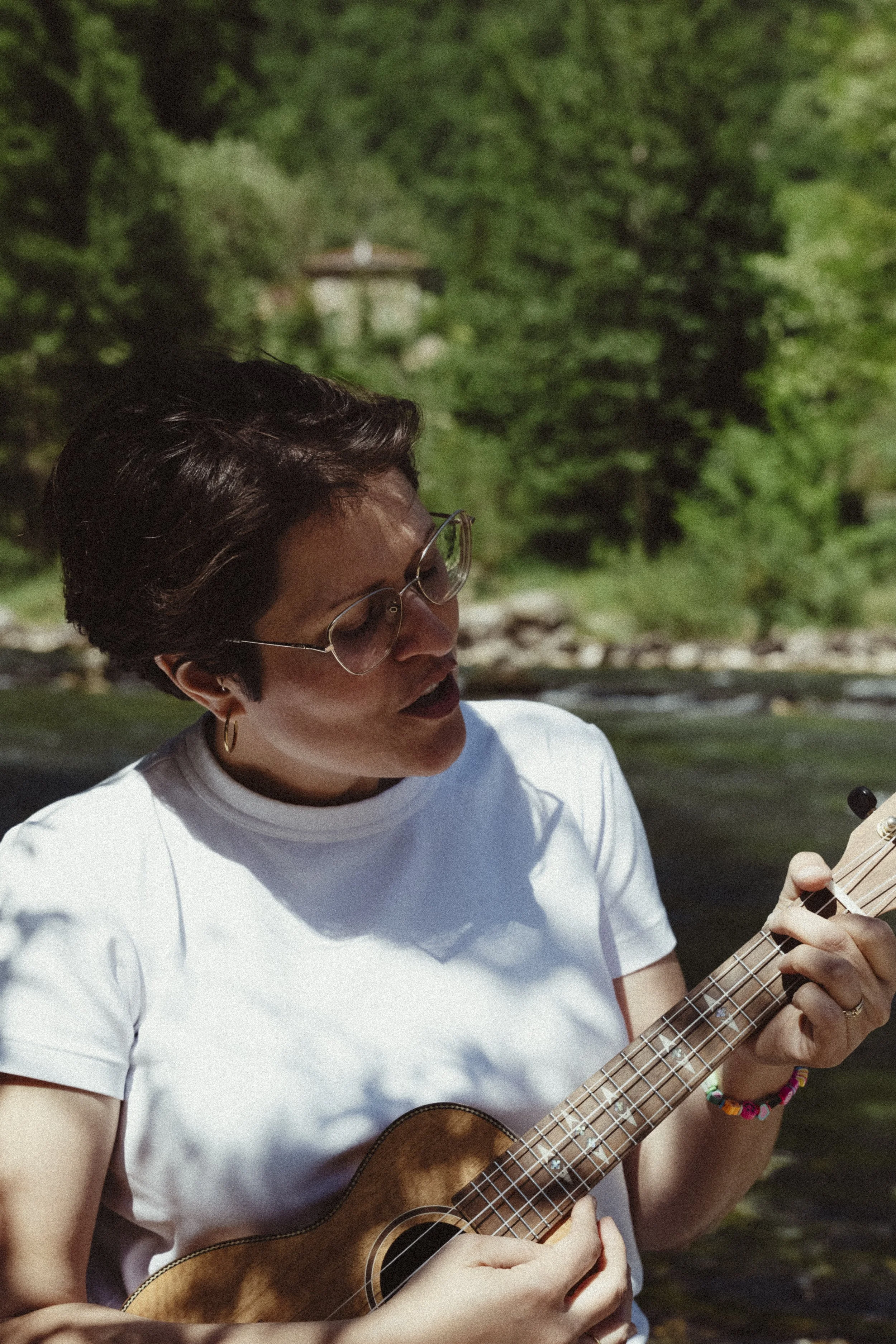 Una donna con gli occhiali che suona una chitarra in un ambiente naturale con alberi e un edificio in lontananza.