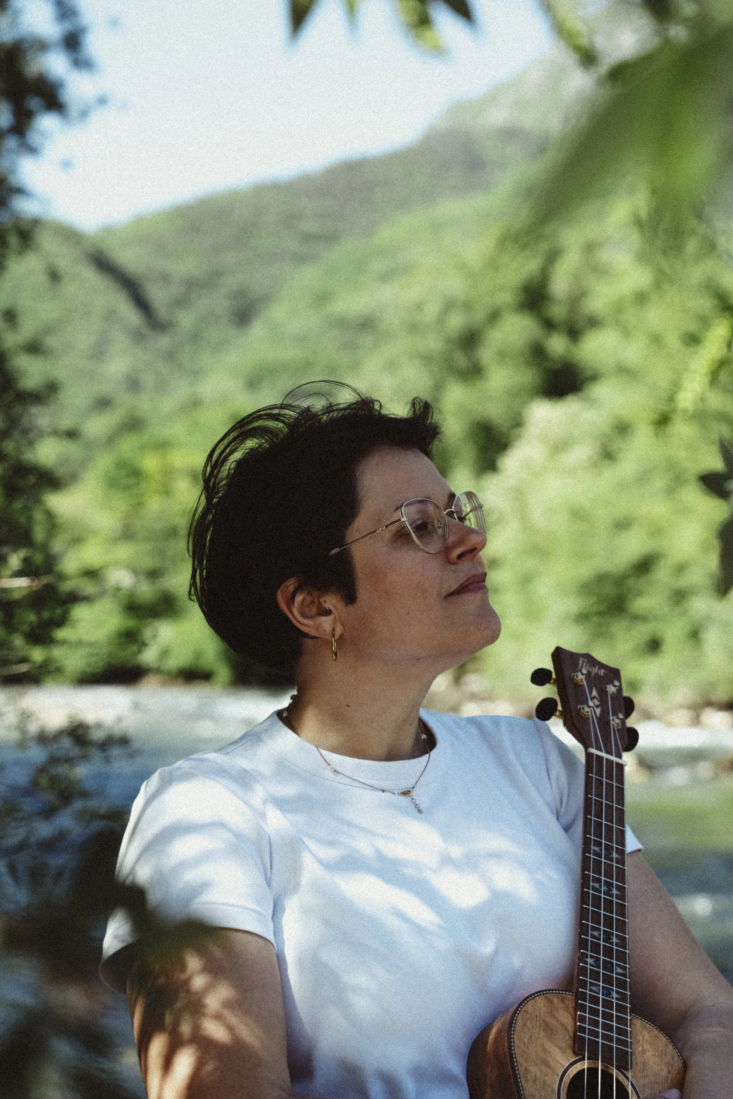 Una donna con capelli corti, occhiali e orecchini d'oro, che suona una chitarra acustica mentre è all'aperto, con uno sfondo di natura verde e un lago.