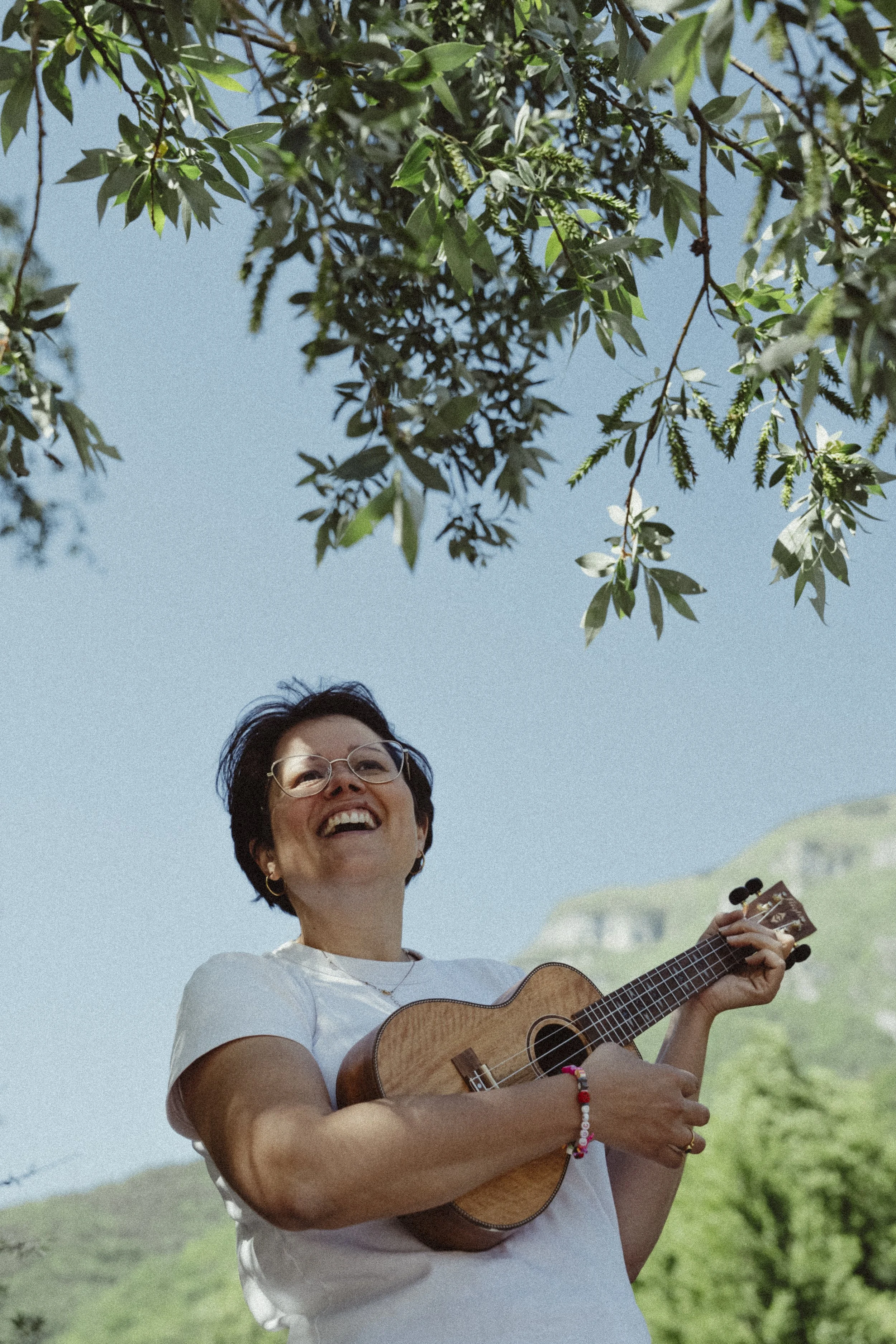 Una donna con occhiali che suona la chitarra sotto un albero, sorridendo con uno sfondo di cielo e colline verdi.