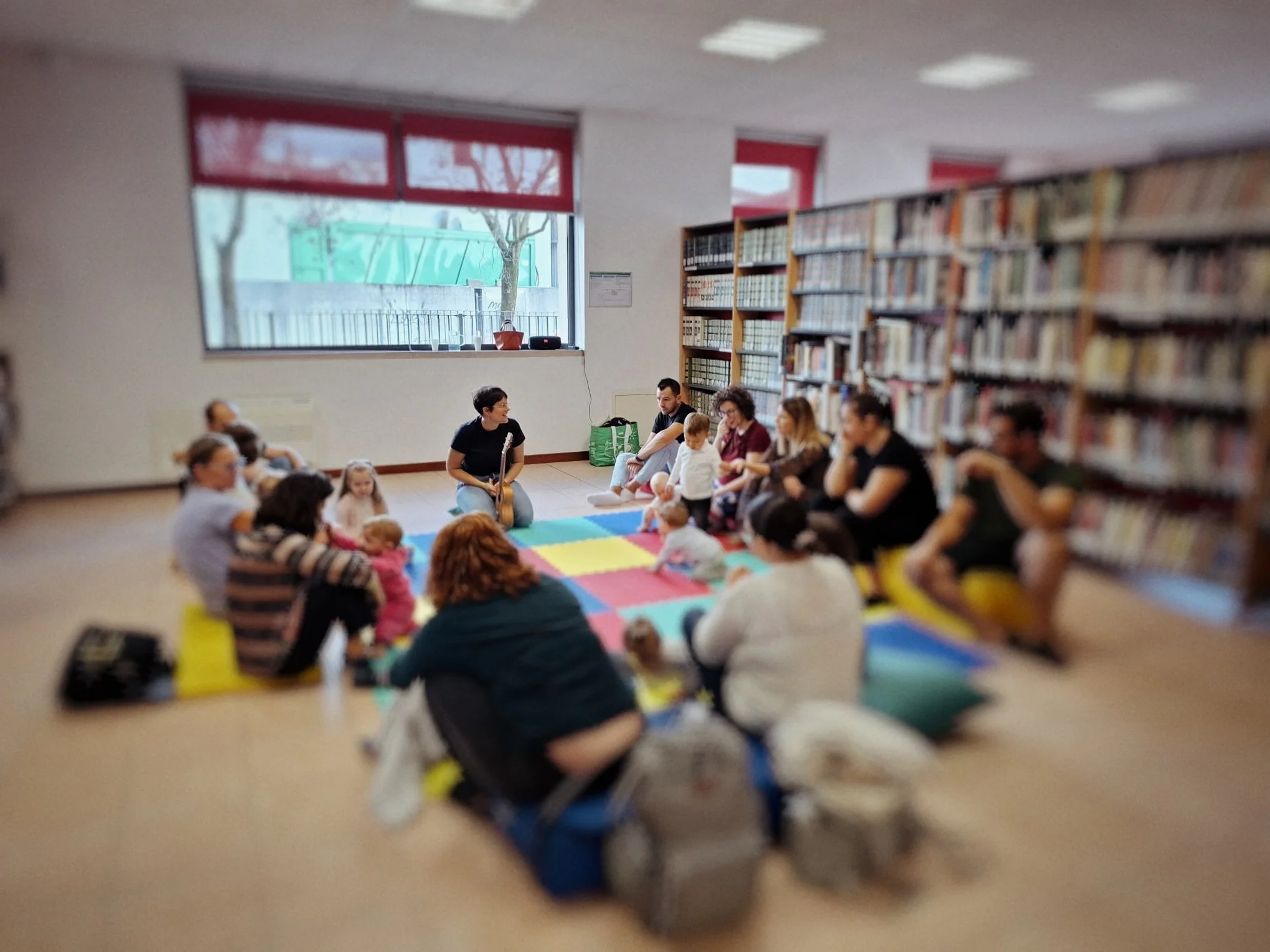 L'immagine mostra un gruppo di persone, tra adulti e bambini, seduti su un tappeto colorato in una biblioteca. Sullo sfondo, scaffali pieni di libri e una finestra con una vista di alberi al di fuori.