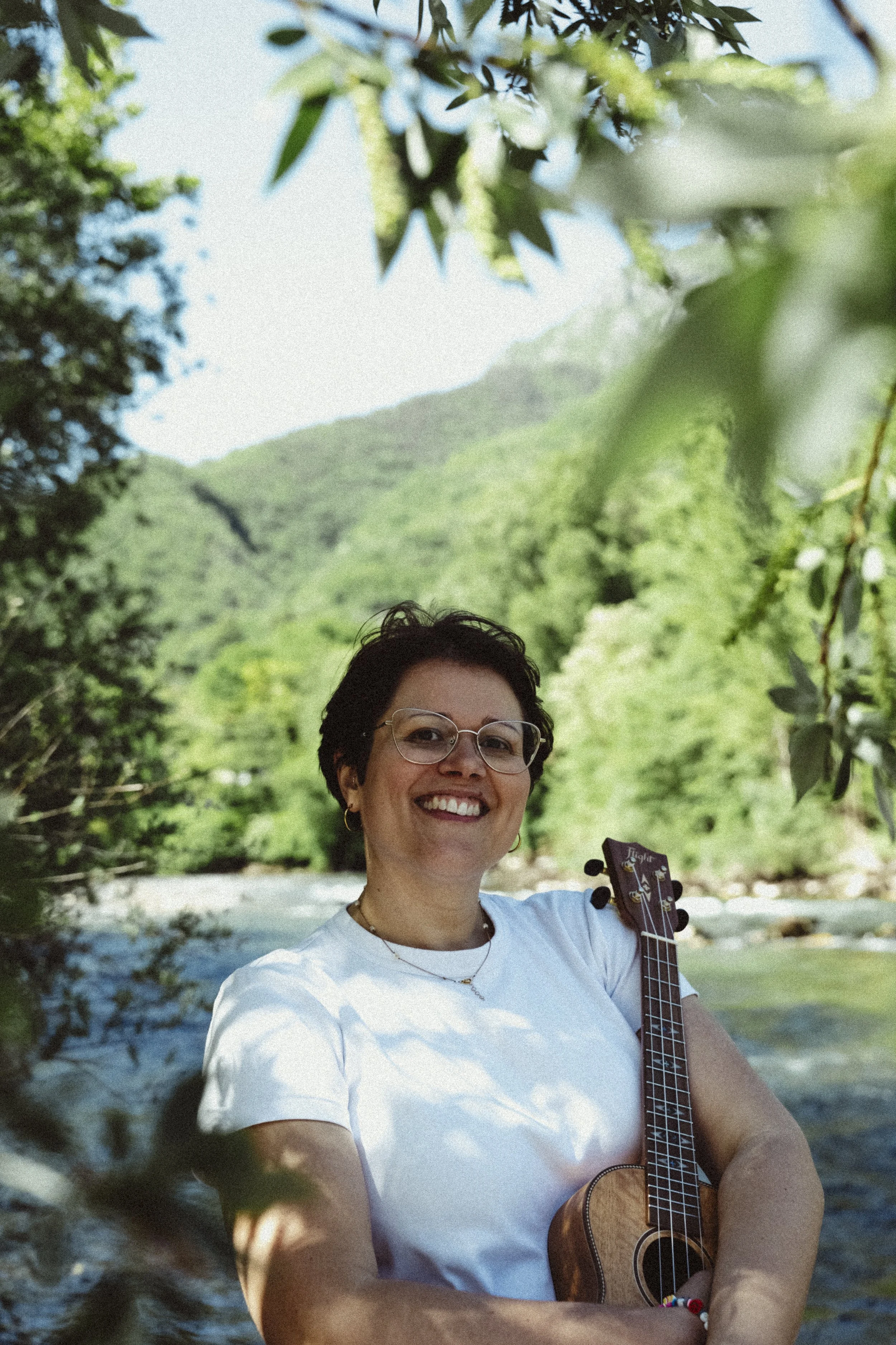 Una donna sorridente con occhiali da vista tenendo una chitarra acustica in un ambiente naturale con alberi, un fiume e montagne sullo sfondo.