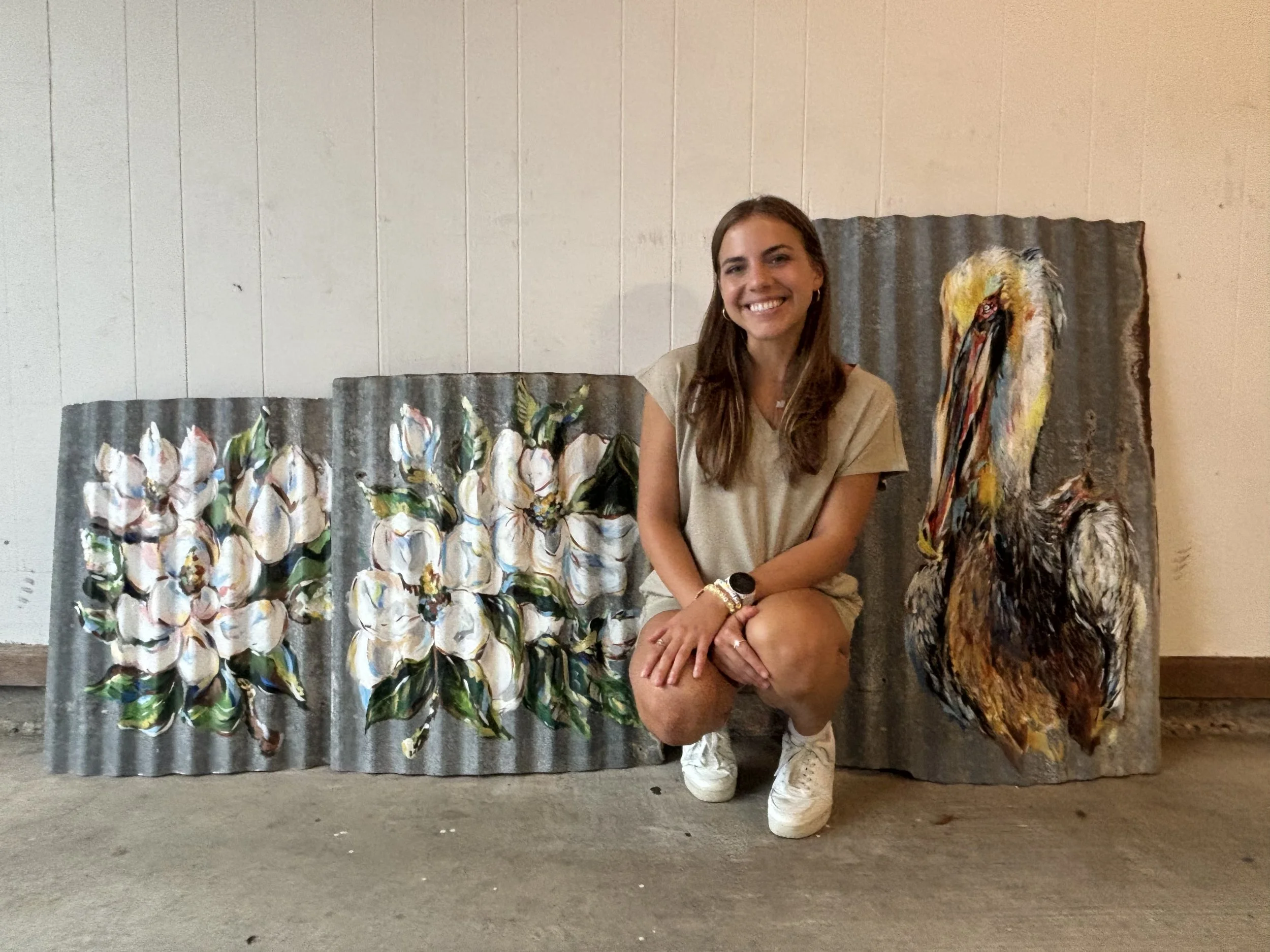 A young woman with long brown hair kneeling on the floor, smiling, wearing a beige shirt, white sneakers, and a watch, in front of three painted corrugated metal panels depicting white roses and a colorful pelican.