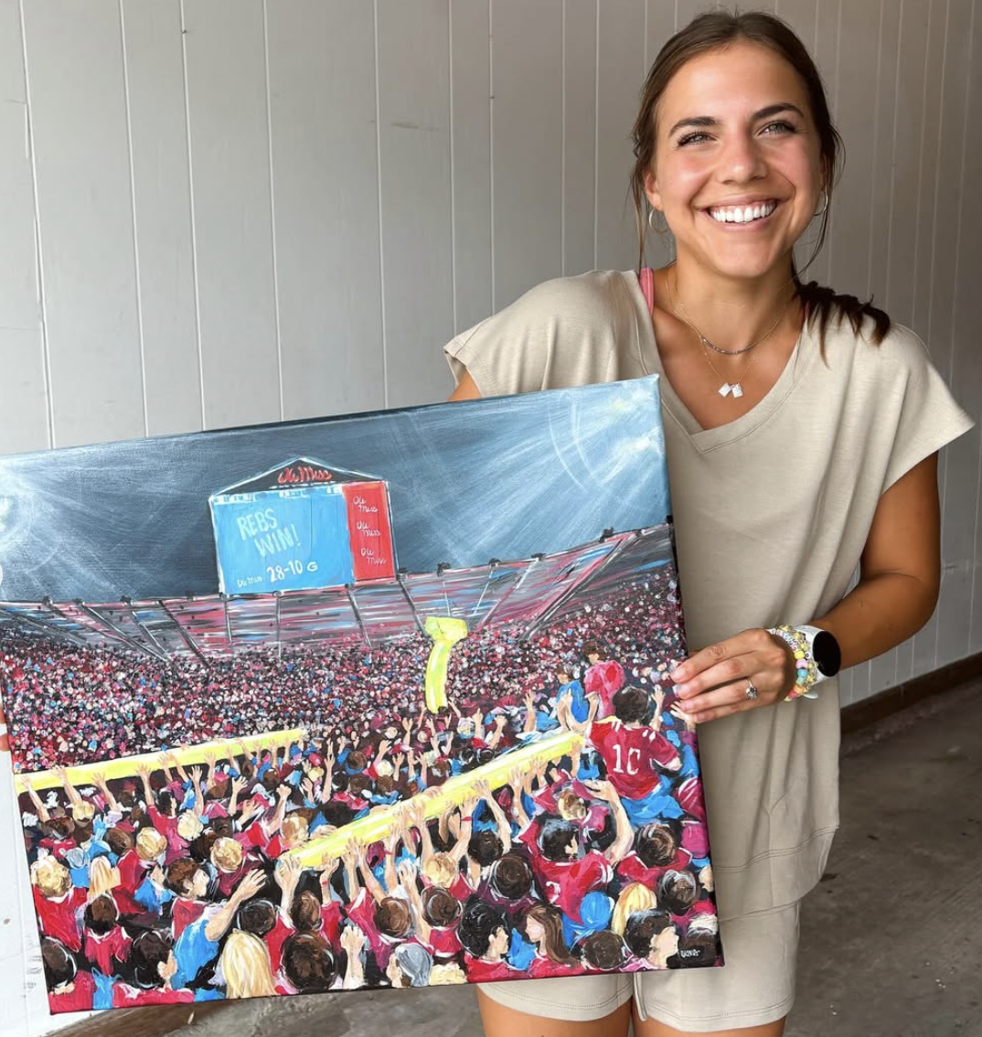 A smiling woman with brown hair holding a colorful painting of a crowded sports stadium scene, with people cheering and a large scoreboard.