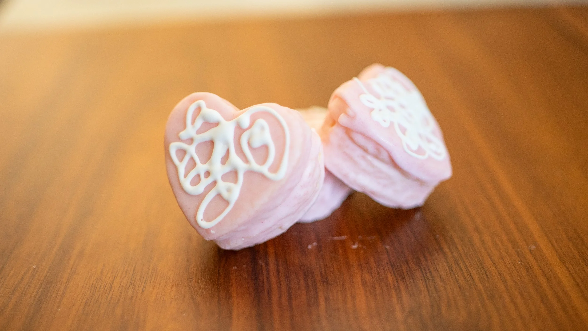 Three pink meringue cookies with white icing decoration on a wooden surface.