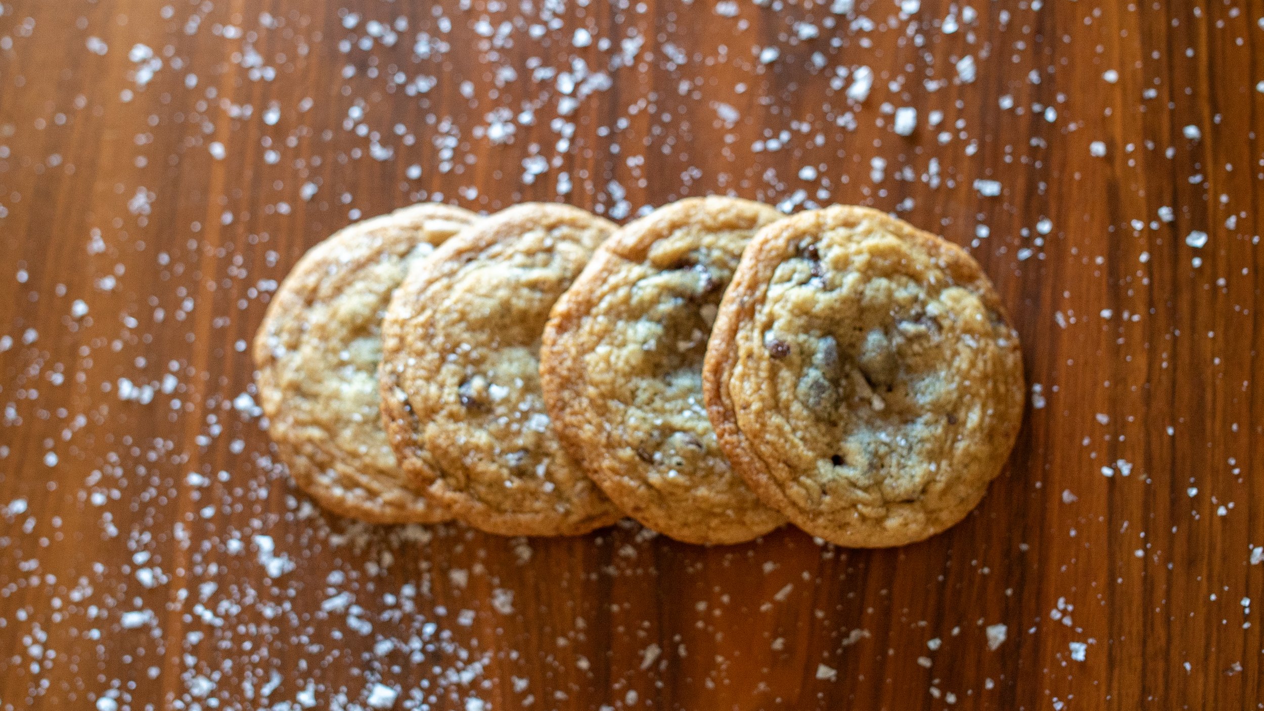 Four chocolate chip cookies sprinkled with powdered sugar on a wooden surface.