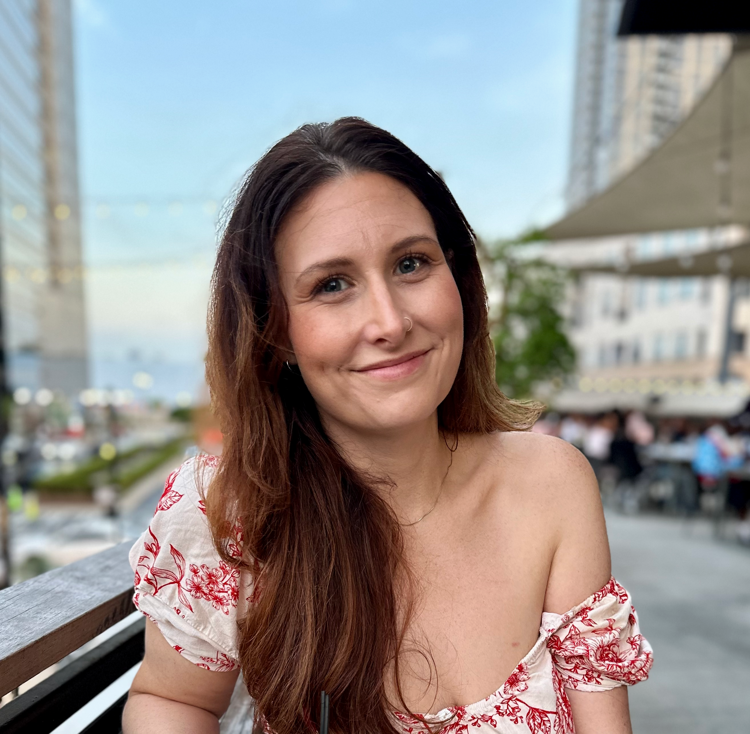 A woman with brown hair, blue eyes, and a nose ring, smiling at the camera outdoors at a restaurant or café with city buildings and a cloudy sky in the background.