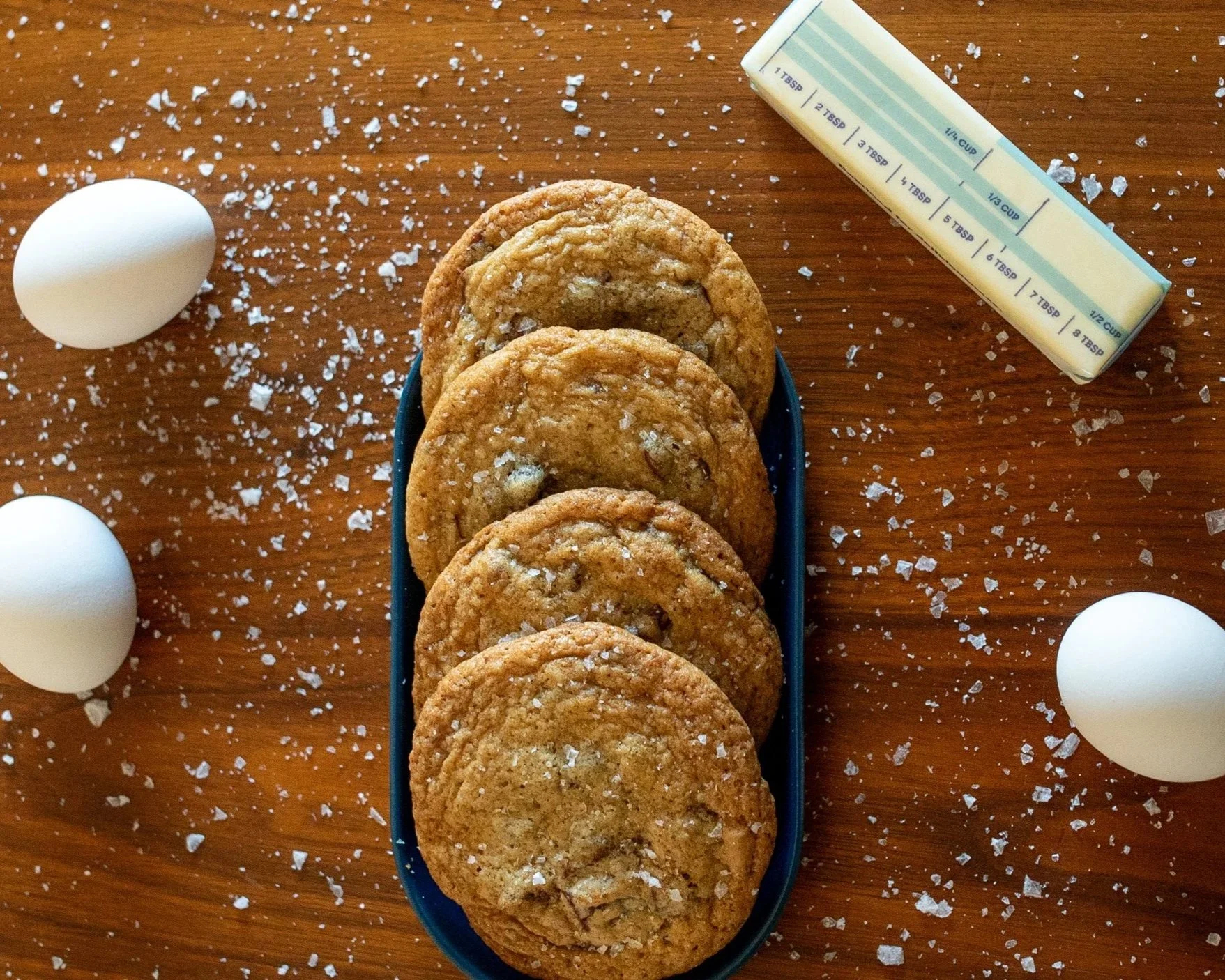 Four chocolate chip cookies in a blue dish on a wooden surface, with a stick of butter, three eggs, and salt scattered around.