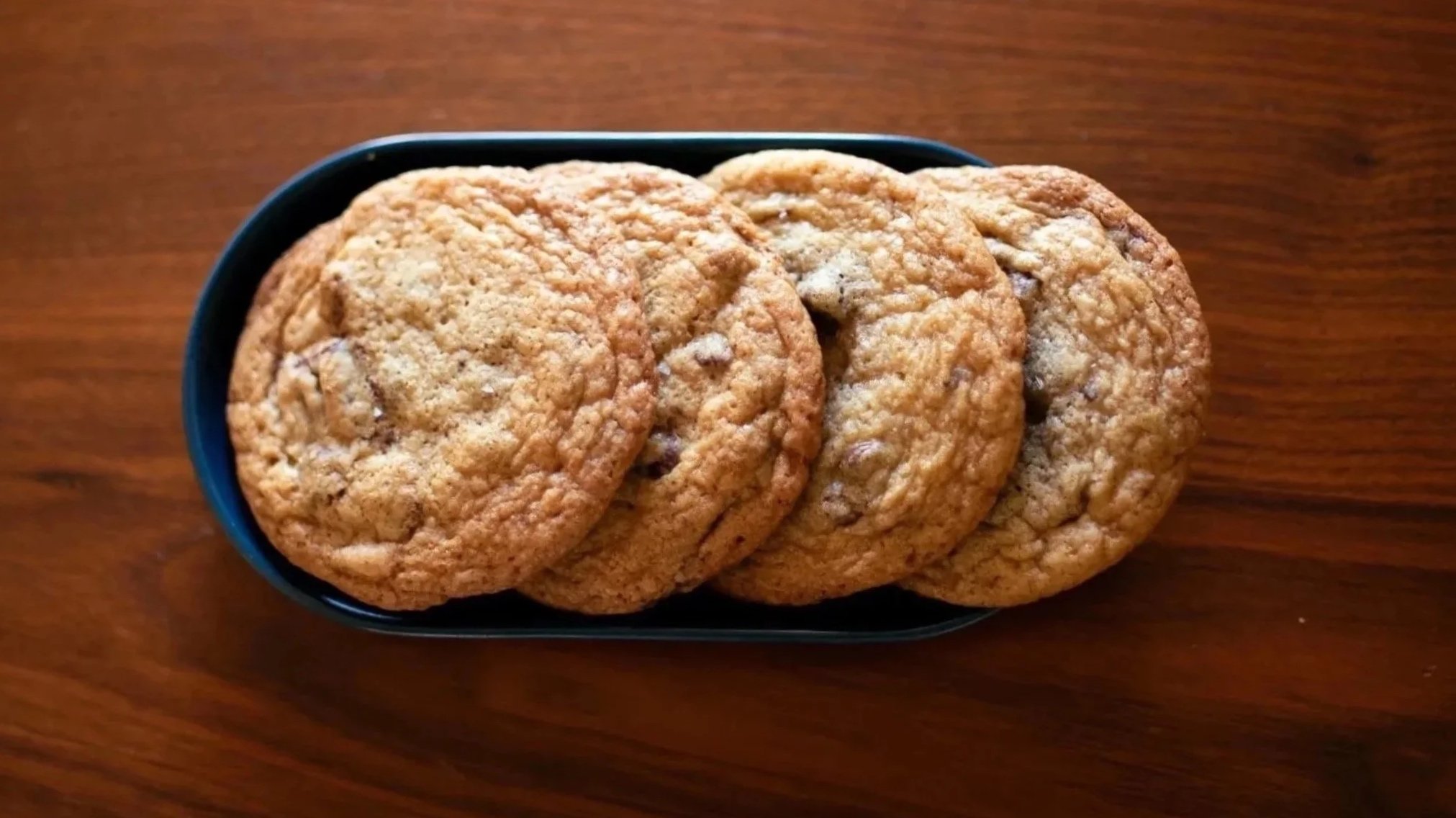 Four chocolate chip cookies on a black oval plate placed on a wooden surface.