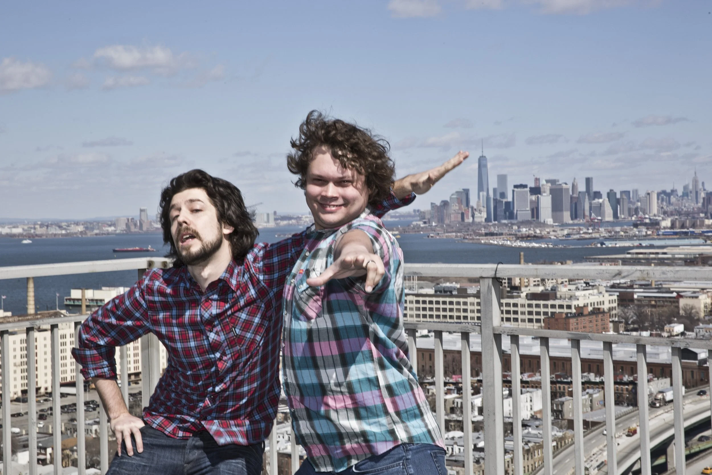 Two young men with curly hair posing on a rooftop with New York City skyline in the background during the day.