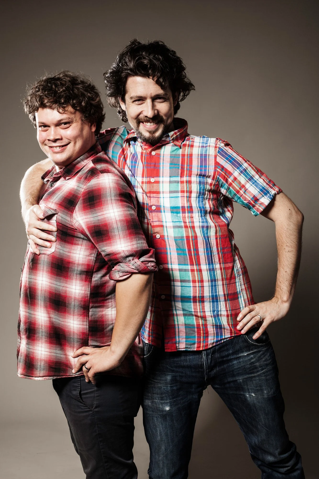 Two men dressed in casual plaid shirts standing close together, smiling, with a neutral background.