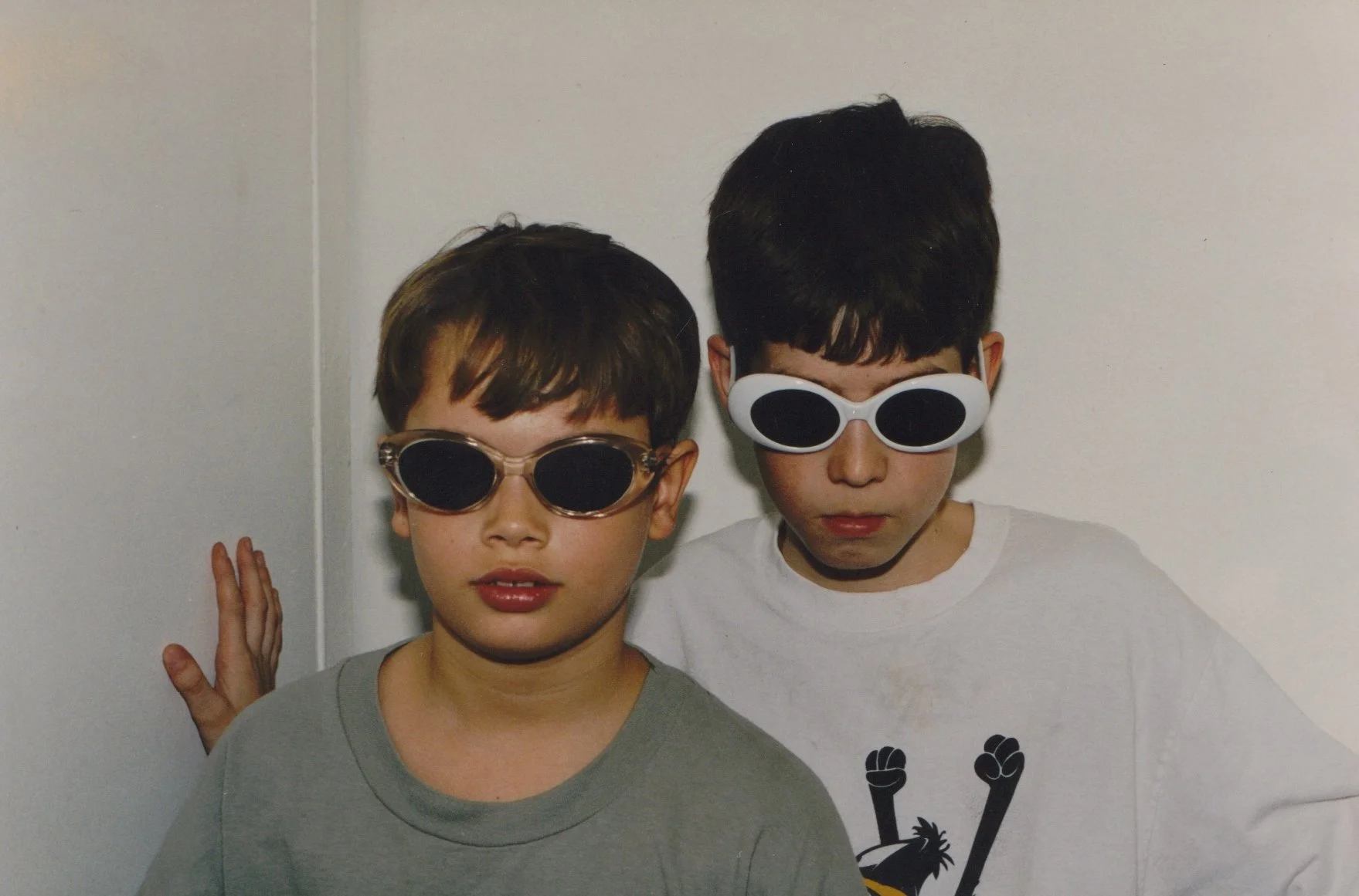 Two boys wearing sunglasses, standing against a plain wall, one with a gray shirt and the other with a white shirt, striking a serious pose.