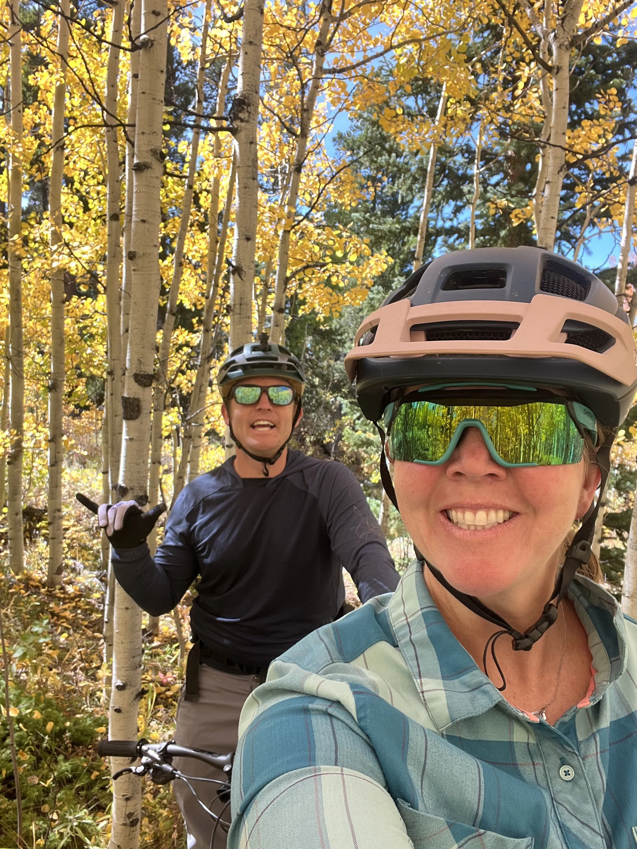Two cyclists, a woman and a man, taking a selfie in a forest with autumn leaves and trees. They are wearing helmets and sunglasses, smiling, and one making a shaka hand sign.