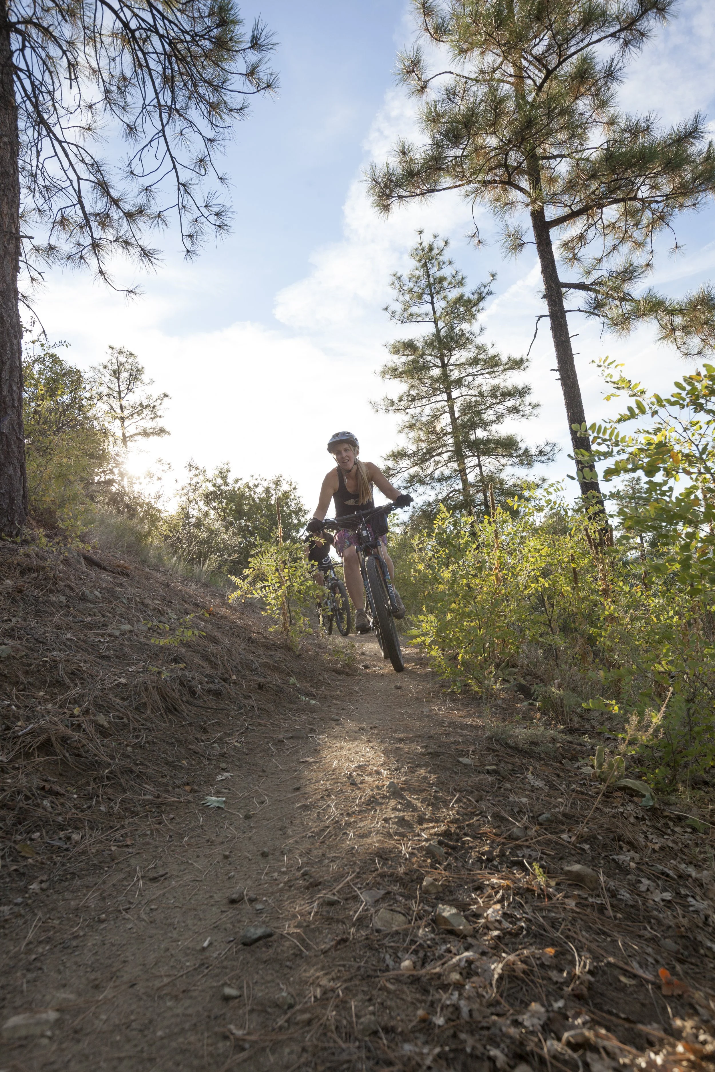 A woman in biking gear, including a helmet and gloves, riding a mountain bike on a dirt trail through a forest with tall pine trees and bushes, under a partly cloudy sky with the sun shining.