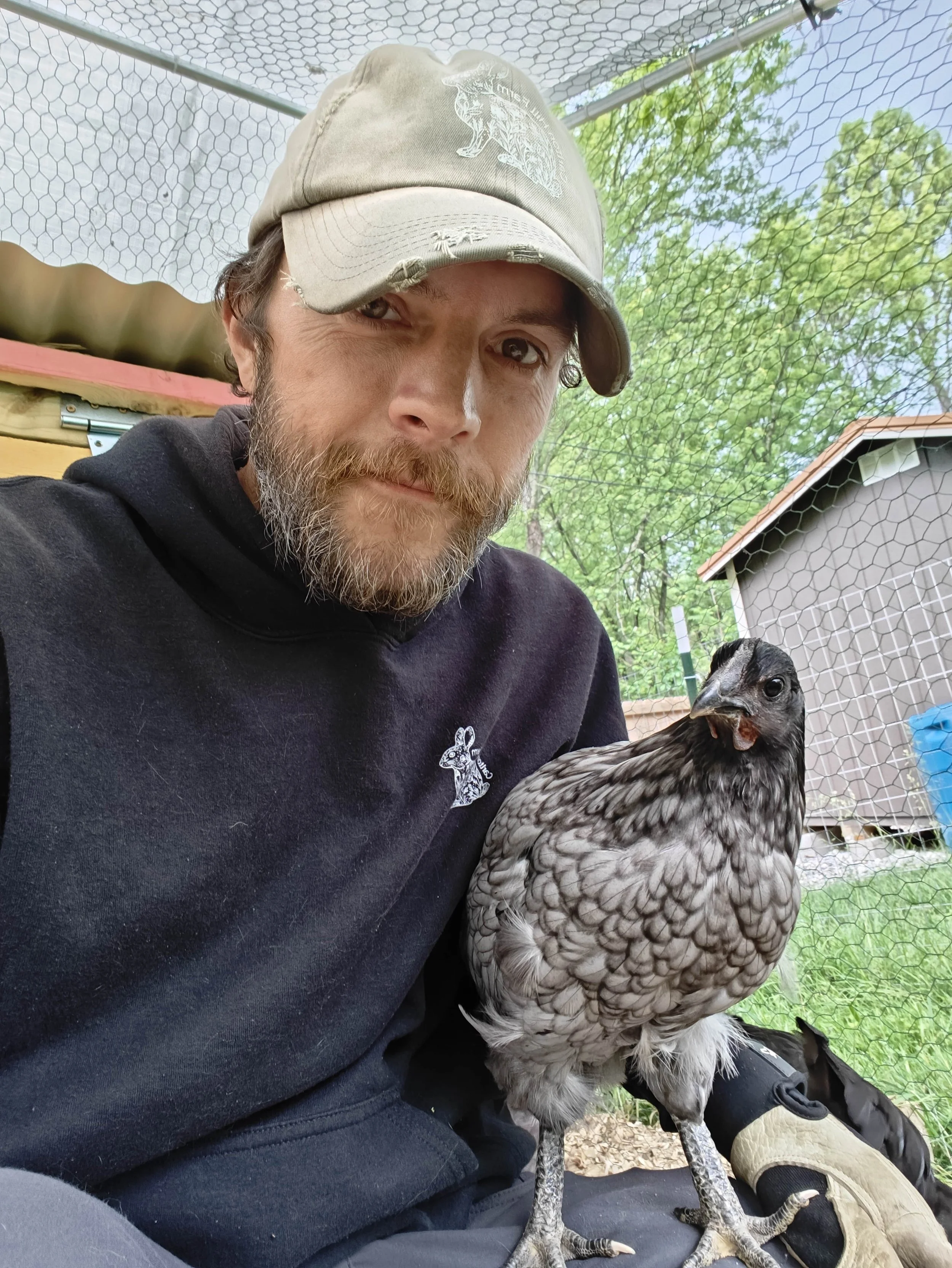 A man with a beard and wearing a beige cap and a black hoodie with a small rabbit logo, taking a selfie with a chicken inside a chicken coop with green trees and a shed in the background.