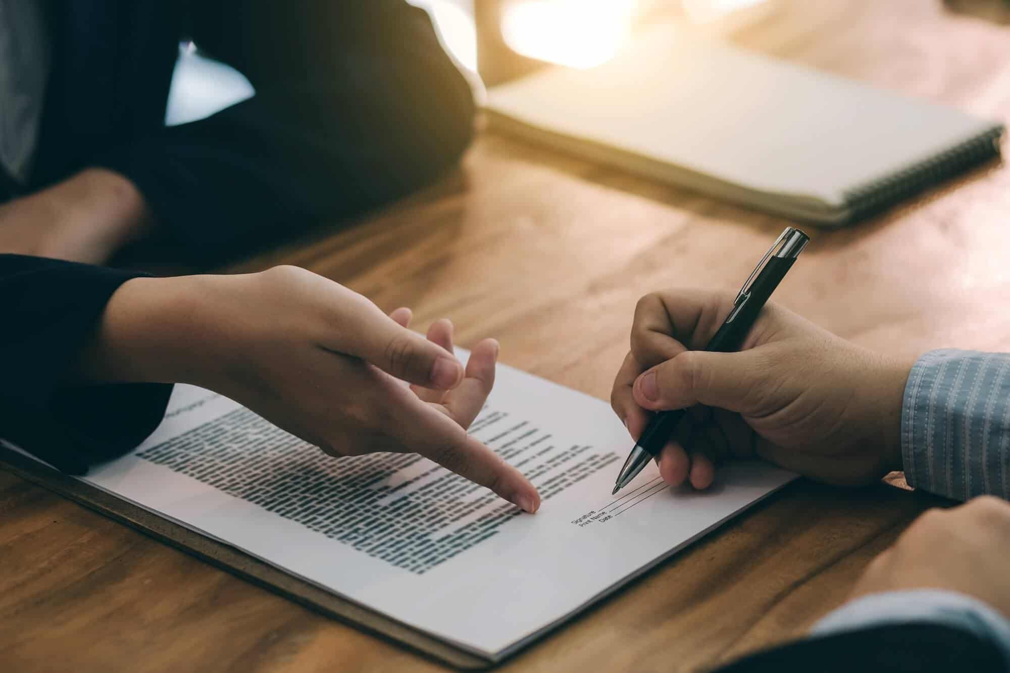 Two people are sitting at a wooden table, one is holding a pen and writing on a paper, the other is pointing at the paper with a finger. There is a notebook in the background.