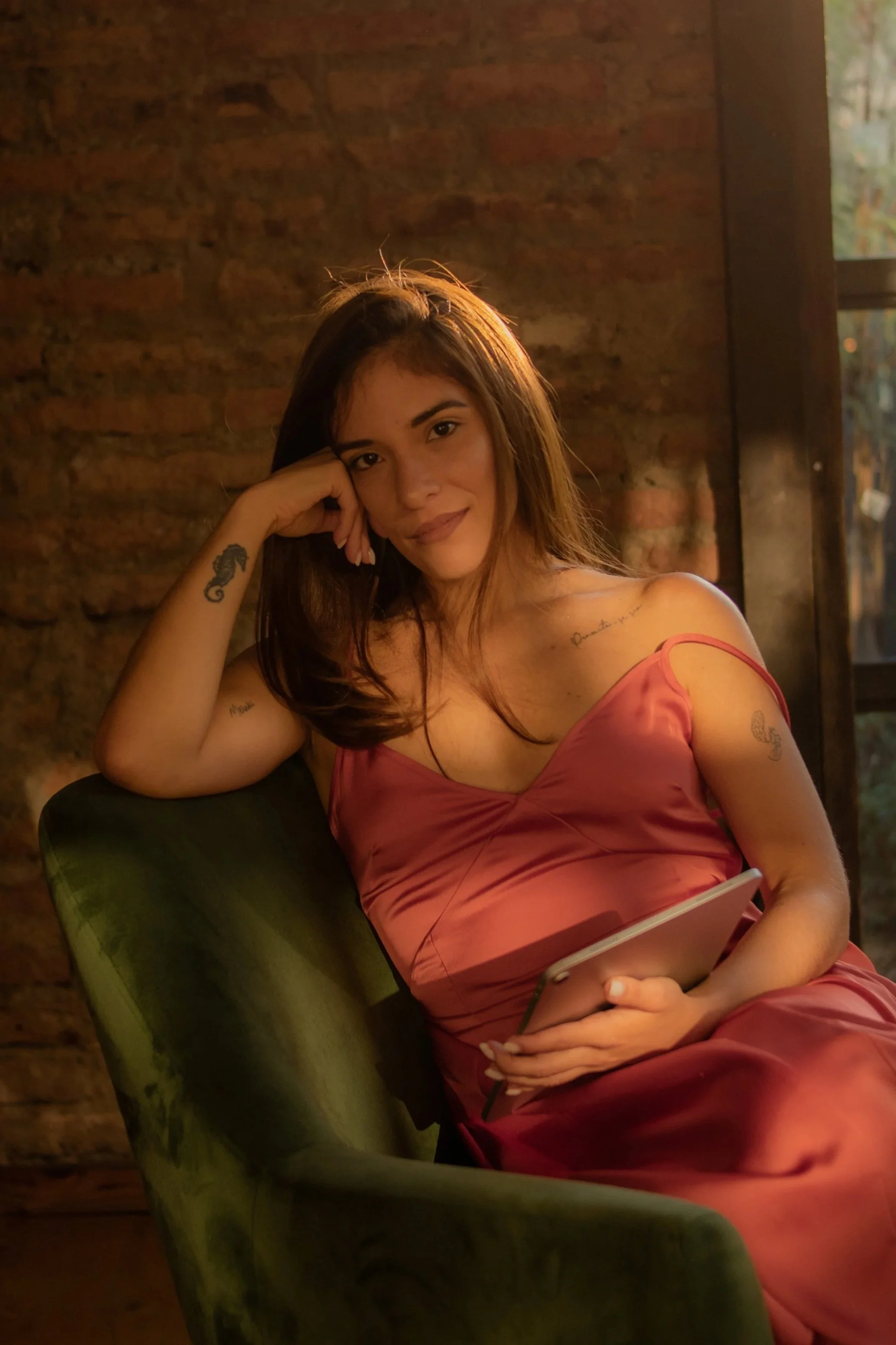 Mujer joven con vestido rosado sentado en una silla, sosteniendo una tableta, en un lugar con pared de ladrillos y luz suave.