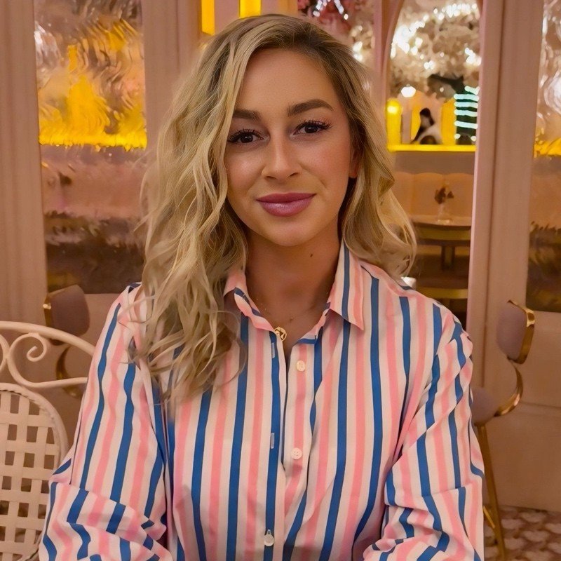 A young woman with blonde wavy hair, wearing a colorful striped button-up shirt, sitting at a table in a warmly lit restaurant or cafe with decorative background and pink floral arrangements.