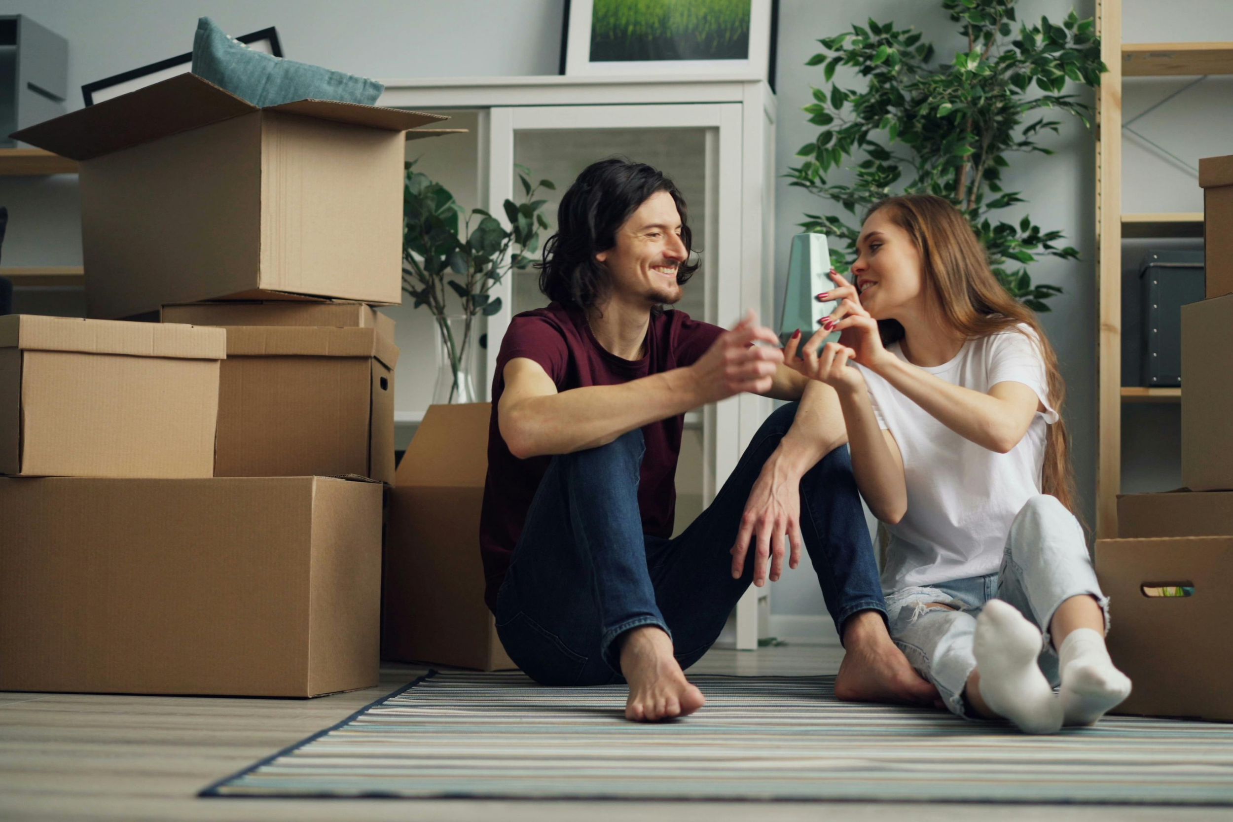A couple sitting on the floor surrounded by moving boxes, smiling and playing with a small dog.