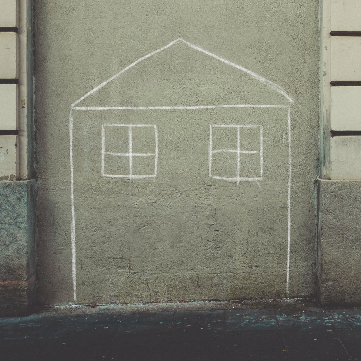 A chalk drawing of a house with two windows on a grey wall.