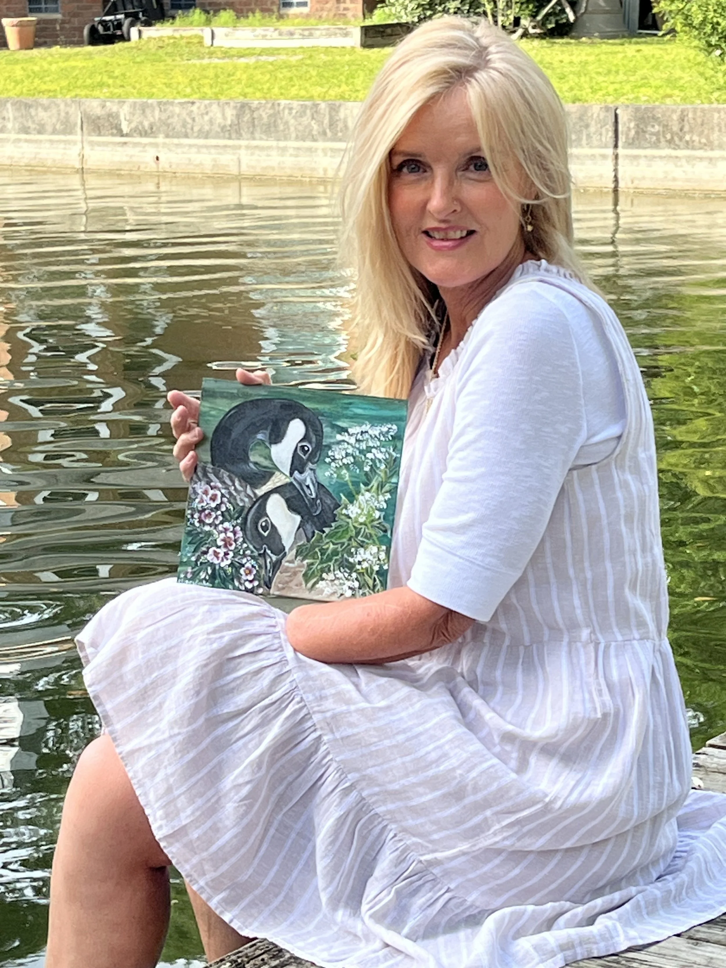 A blonde woman sitting on a dock by a lake, holding a painting of two black and white ducks surrounded by flowers and greenery.