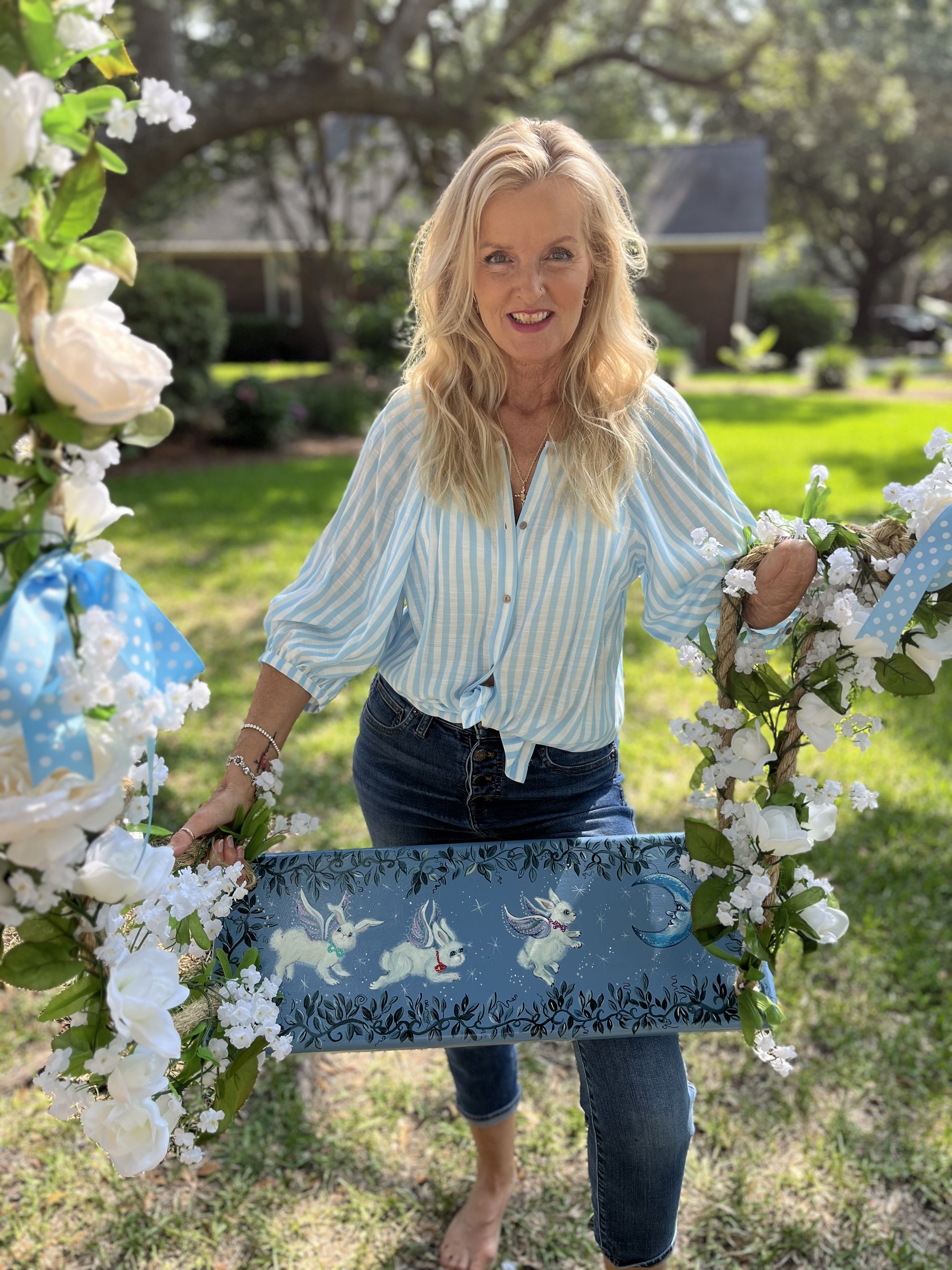 A woman with blonde hair smiling, holding a decorated wreath or sign with white bunny illustrations and a blue background, outdoors on a sunny day with green grass and trees in the background.