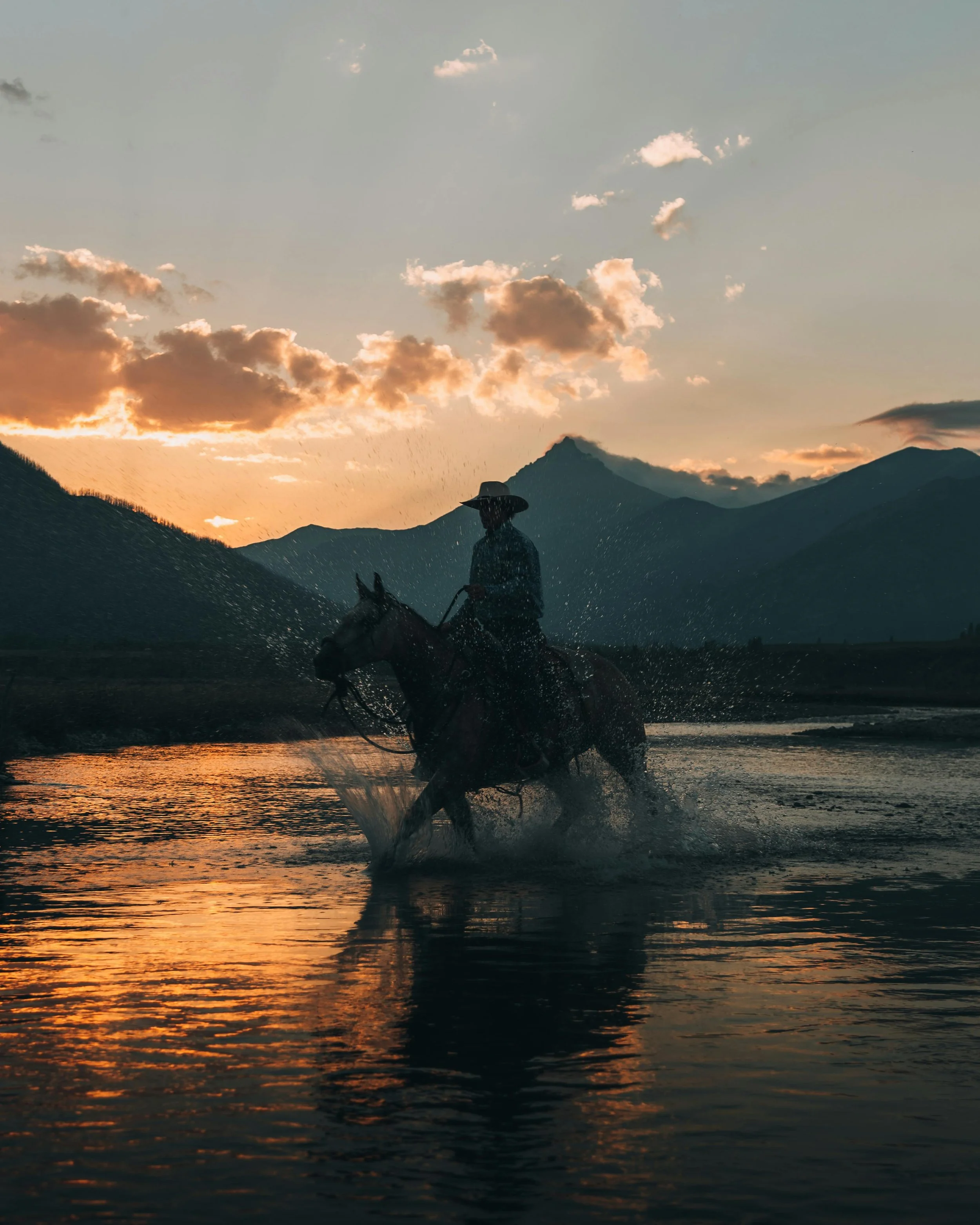 A person riding a horse through water at sunset, with mountains and clouds in the background.
