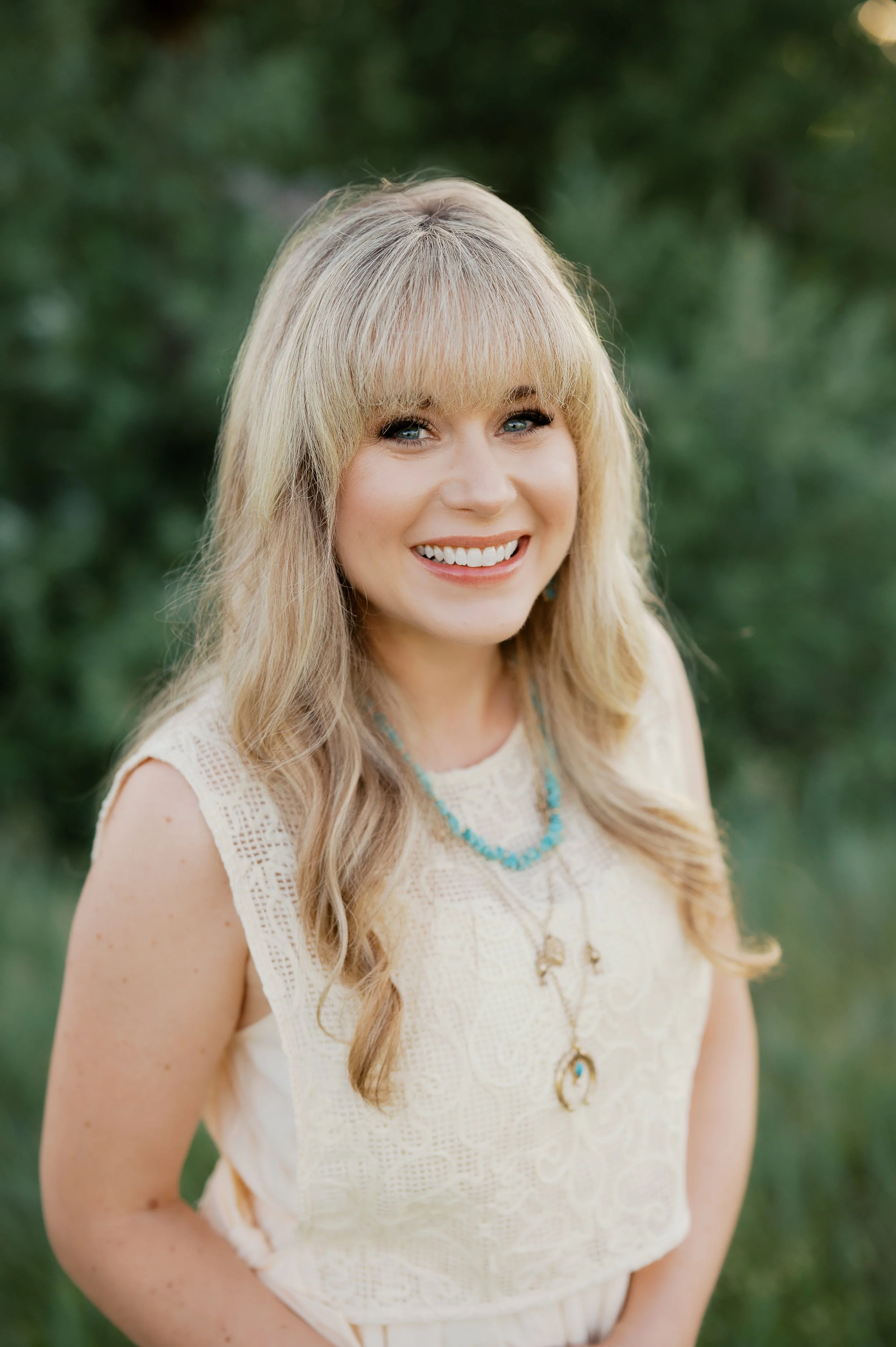 A woman with long blonde hair and blue eyes smiling outdoors with a green blurred background, wearing a cream-colored sleeveless lace dress and turquoise jewelry.