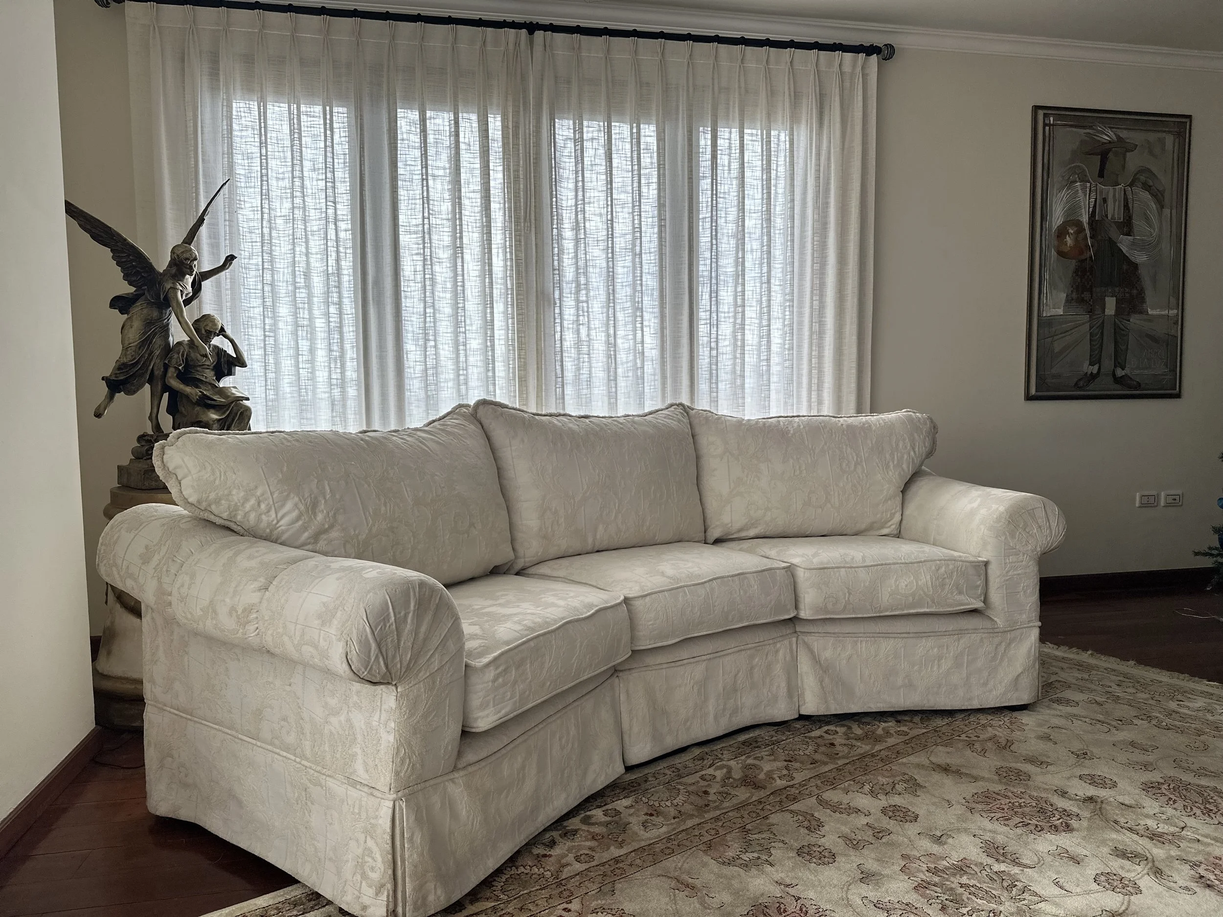 Living room with a beige sofa, a chandelier with a sculpture of angels, window with white curtains, framed artwork on the wall, area rug, and hardwood flooring.
