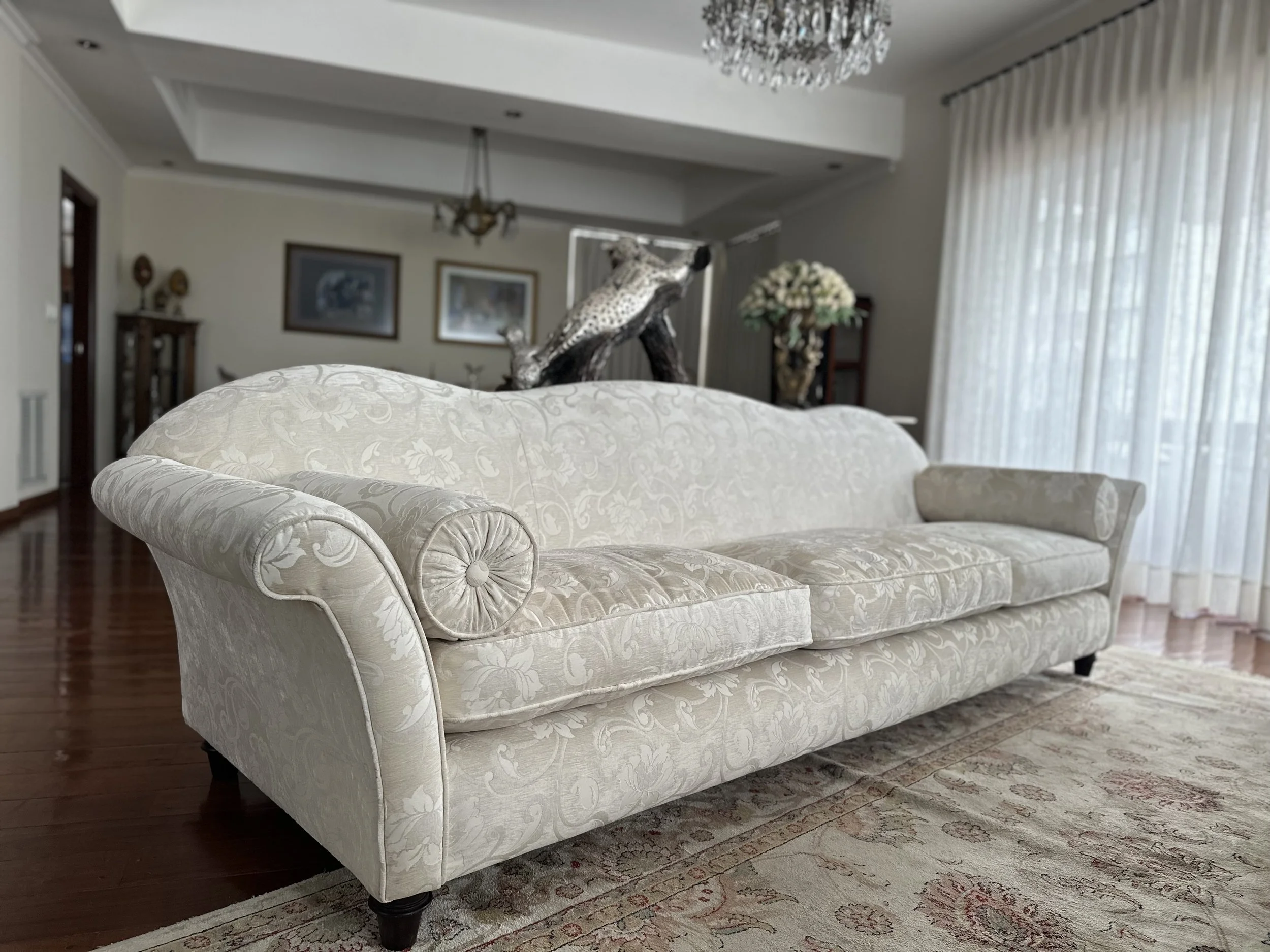 A cream-colored, floral-patterned sofa with rolled arms and dark wooden legs in a living room with hardwood floors, curtains, and decorative items.