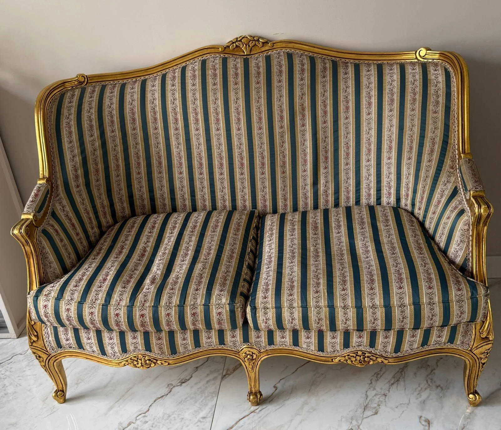 Ornate vintage sofa with gold frame and striped fabric upholstery in blue, gold, and beige, placed on a marble floor.