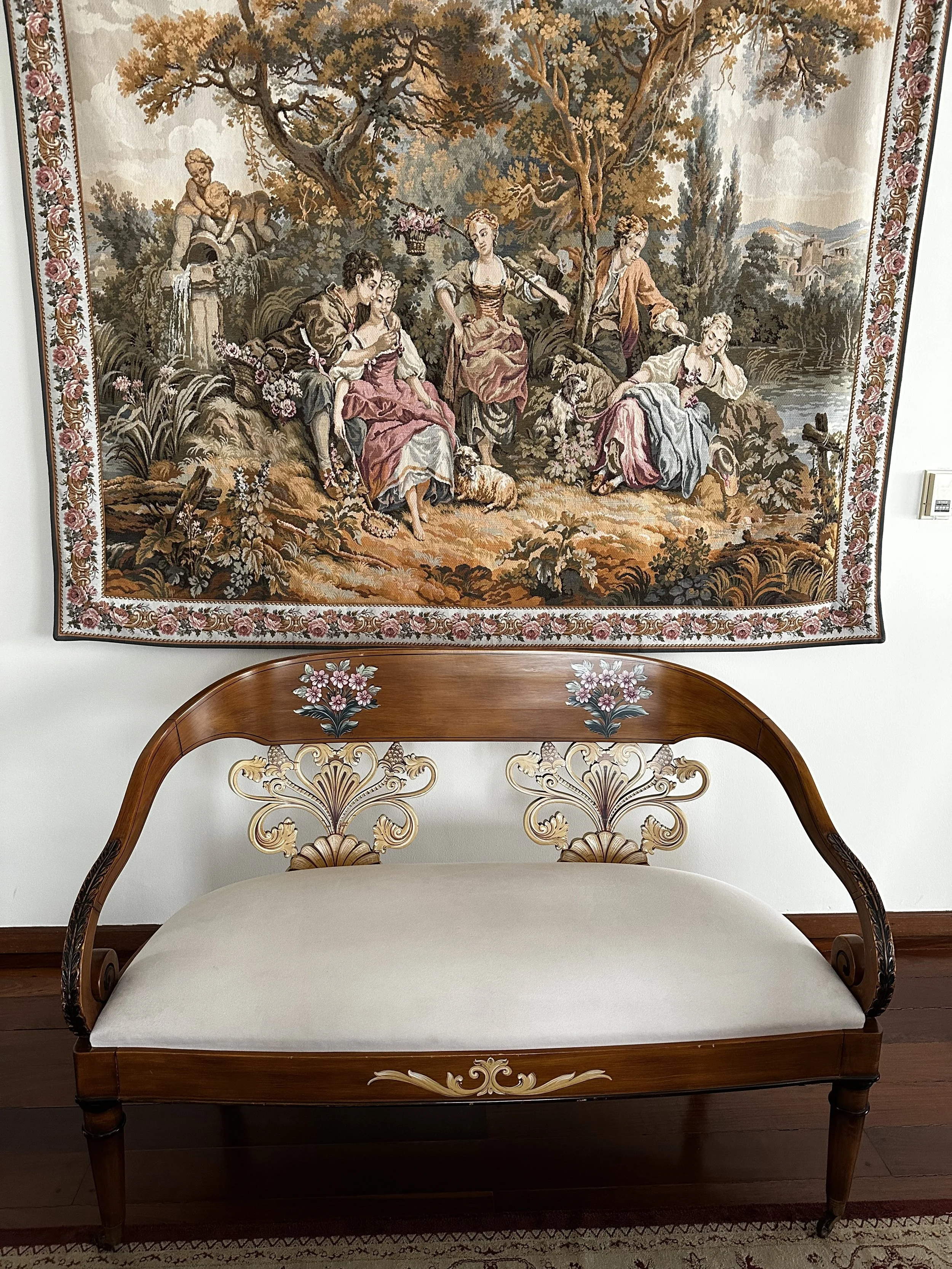 A tapestry hung on a wall featuring a classical pastoral scene with several women and men in an outdoor setting near a river, surrounded by trees and flowers. Below the tapestry, there is an ornate wooden bench with floral carvings and inlays, and a 