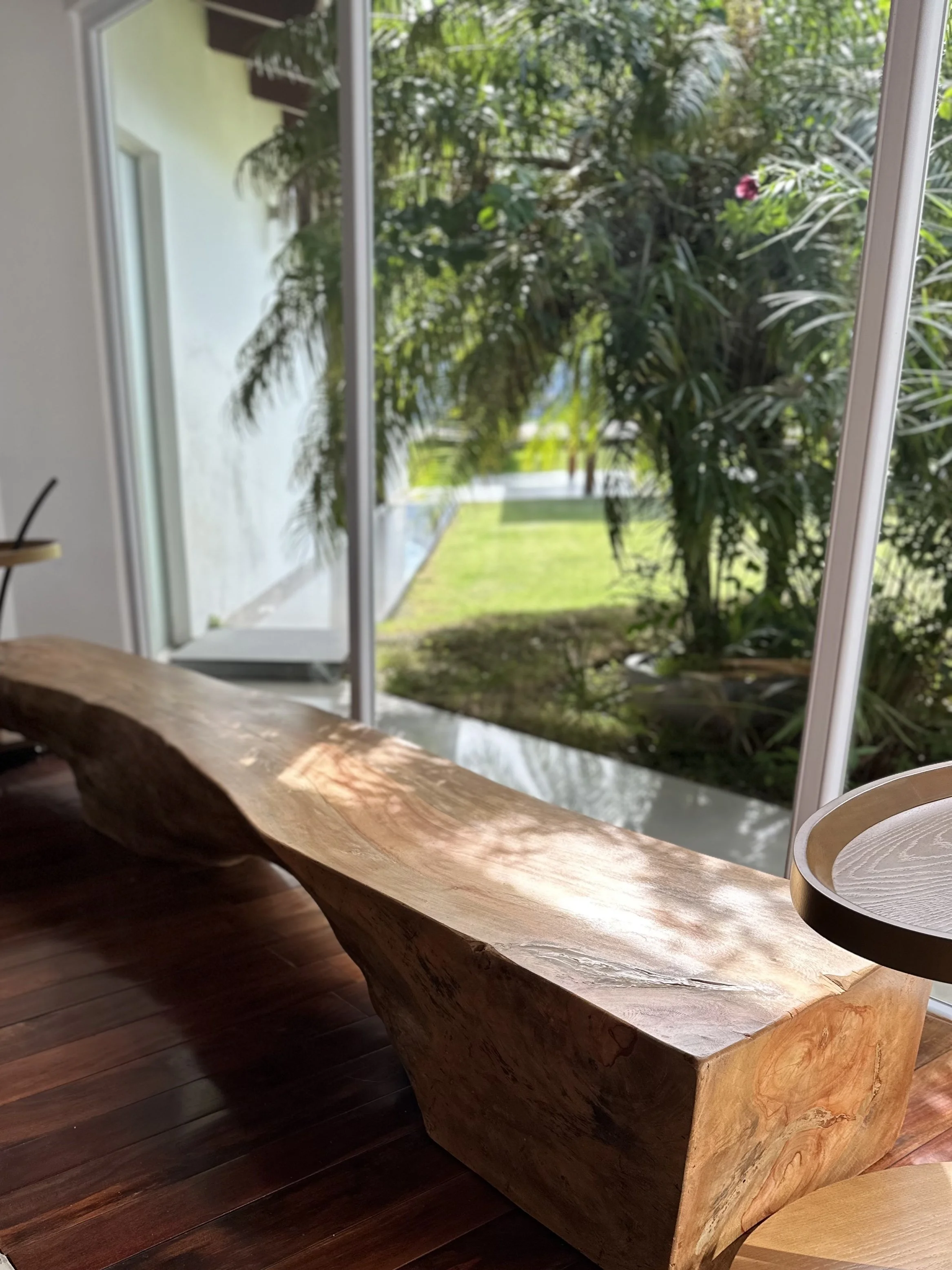 Interior with a large carved wooden bench placed near floor-to-ceiling windows showing green garden outside.