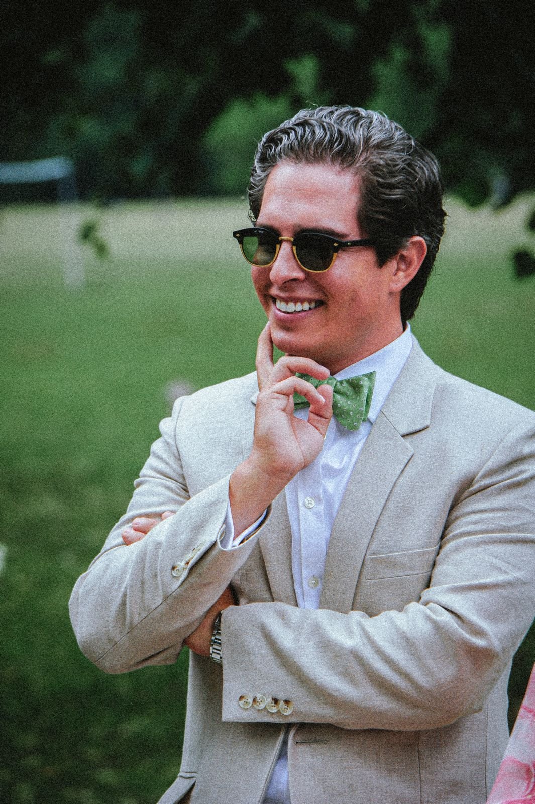 A man with dark, wavy hair wearing sunglasses, a light-colored suit, a white shirt, and a green polka-dotted bow tie, standing outdoors on a grassy area with trees in the background, smiling thoughtfully with his hand on his chin.
