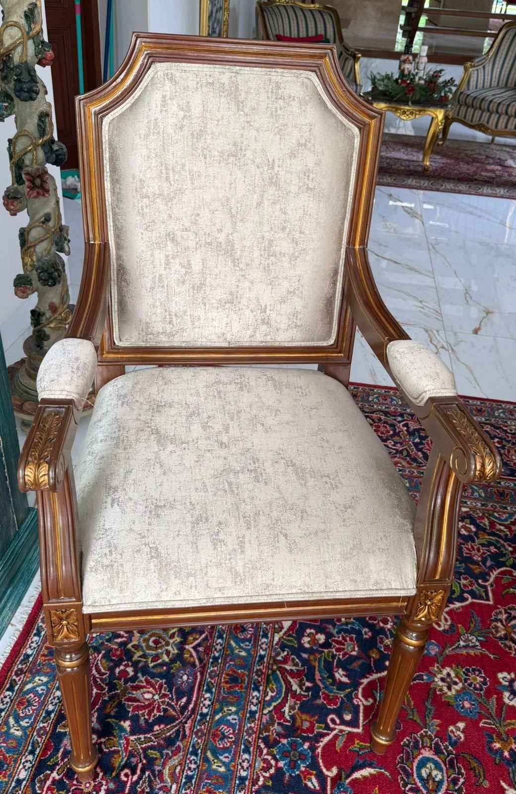A vintage beige upholstered armchair with carved wooden frame and armrests, placed on a colorful patterned rug in a room with other antique furniture and marble flooring.